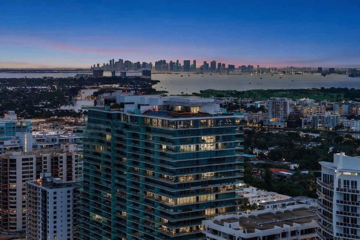 This aerial view showcases a modern high-rise building with a sleek, glass-clad facade, offering stunning panoramic views of the city skyline and waterfront. The building features multiple balconies and a rooftop amenity area, suggesting luxurious living. The surrounding area includes lush greenery and other residential buildings, creating a vibrant urban landscape.