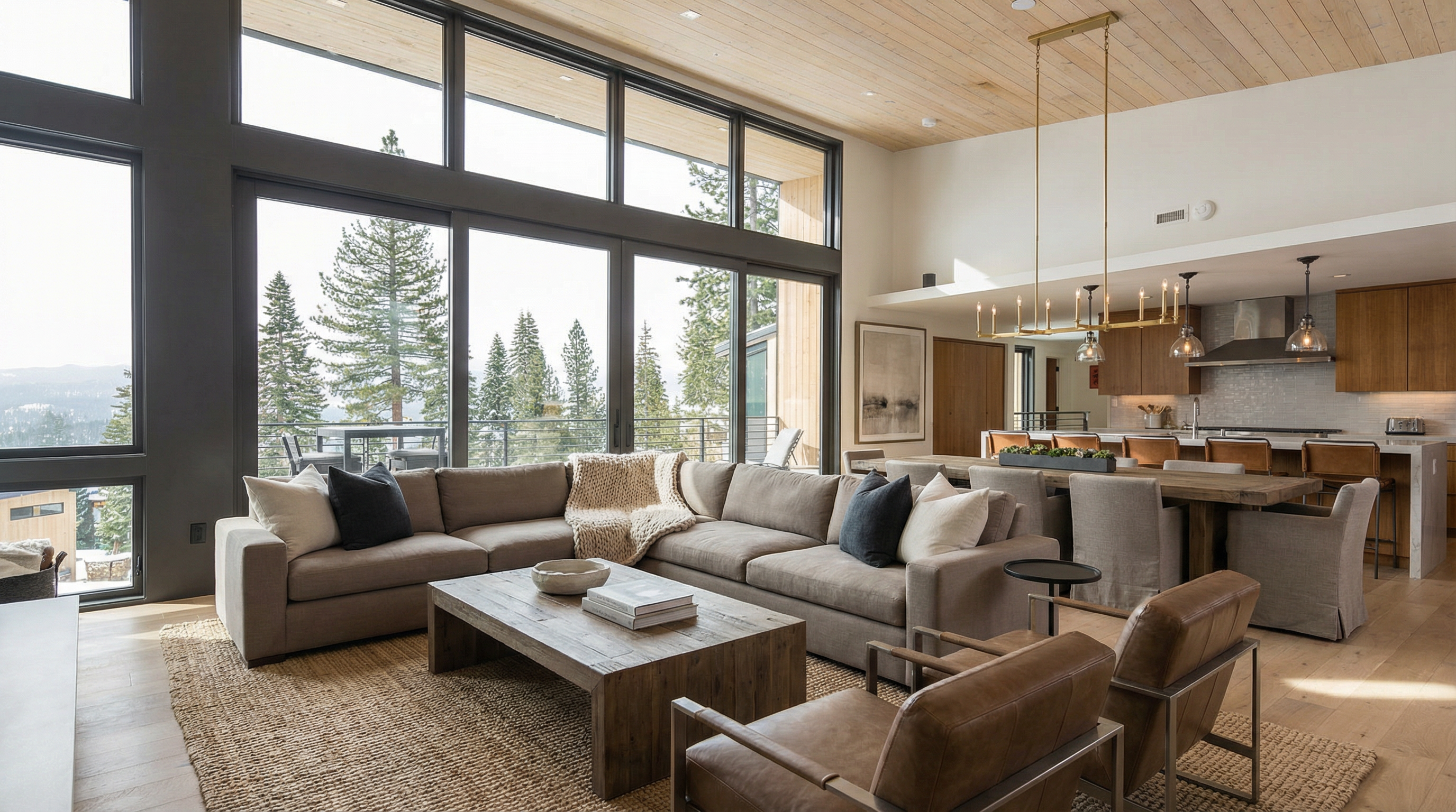 This is a spacious living room featuring a large sectional sofa, a leather ottoman, and a chaise lounge. The room has floor-to-ceiling windows that offer a view of the snow-covered trees outside. The open floor plan connects the living room to the kitchen and dining area, creating a seamless flow for entertaining.