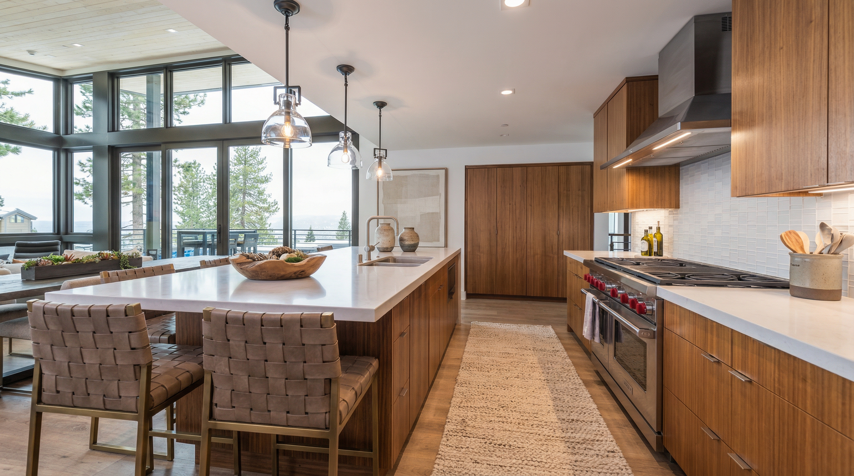 This is a bright and modern kitchen featuring wood cabinetry, a large island with a white countertop, and stainless steel appliances. Large windows offer a view of the outdoors, and pendant lights hang above the island. The kitchen has a clean and contemporary design with a focus on natural light and open space.