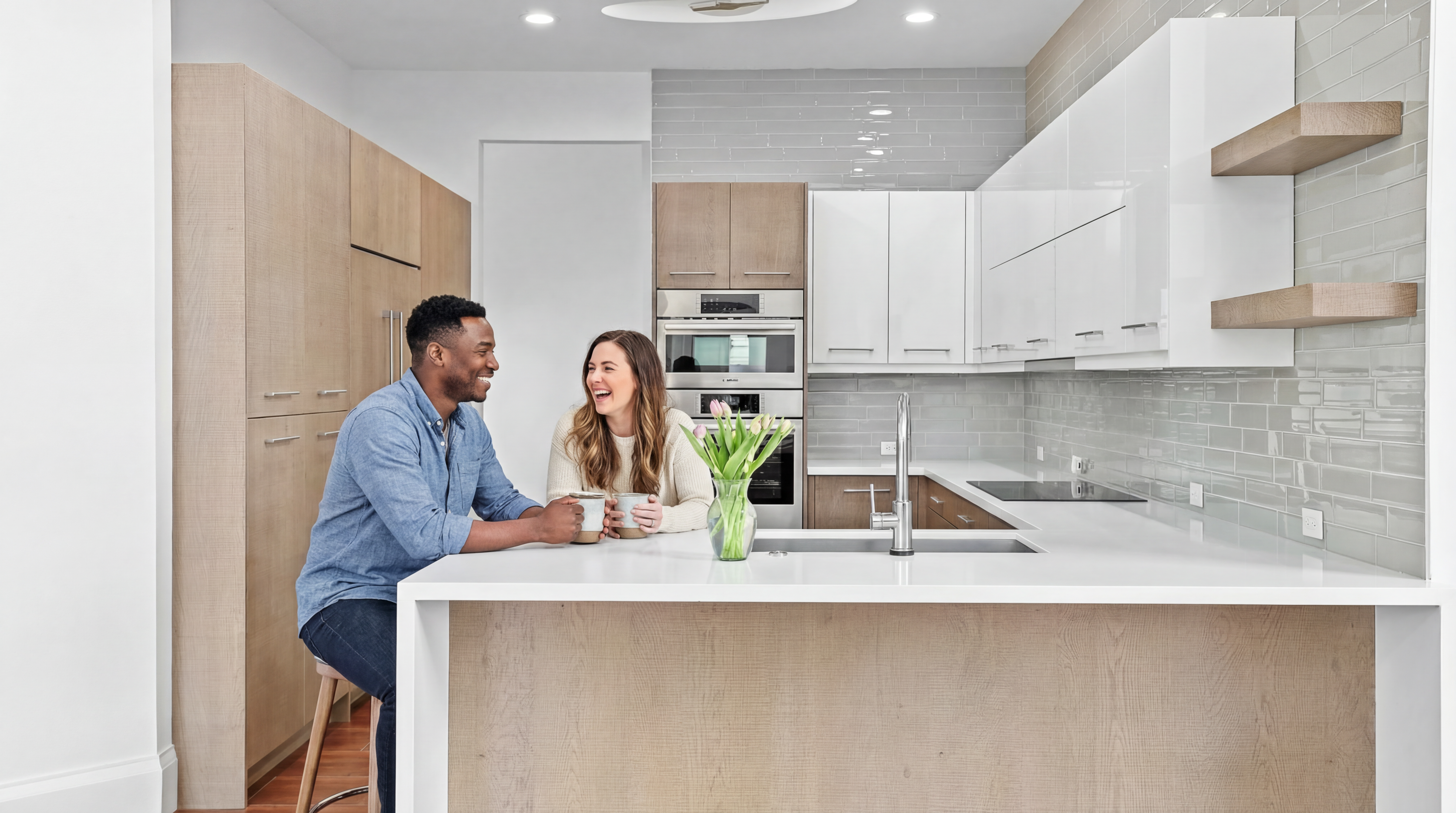 This is a well-lit, modern kitchen featuring a large island with a white countertop and light wood paneling. The kitchen includes stainless steel appliances, a combination of white and wood cabinetry, and a light gray backsplash. The flooring appears to be a reddish-brown tile, and a vase of flowers sits on the island, adding a touch of color.