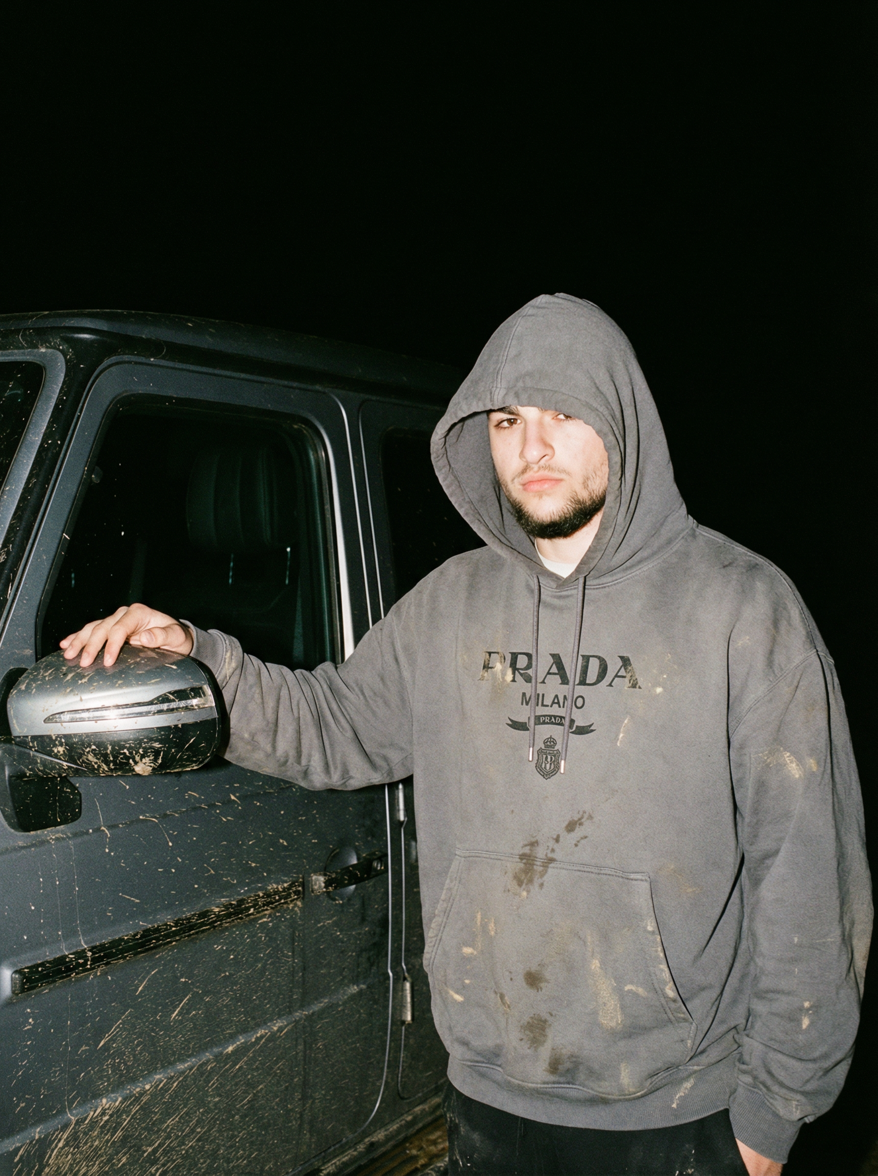 A hyper-realistic nighttime full-body photograph of a person wearing an oversized Prada logo muddy hoodie in gray, standing beside a muddy dark Mercedes G-Wagon, the Prada logo slightly distorted by fabric folds and creasing naturally across the chest, hood partially up casting shadow over the eyes, one hand resting on the car's side mirror, direct on-camera flash creating sharp highlights on the hoodie's fabric surface and the cars paint, deep shadows behind both subject and vehicle falling to black void, cinematic grain, visible fabric texture showing the weight and pilling of the cotton, the logo catching flash unevenly due to fabric deformation, tactile detail on the car's door handle showing fingerprints, documentary fashion photography, intimate and unposed feeling, shot on 35mm film style, shallow depth of field, rawrealism | User Input: N/A