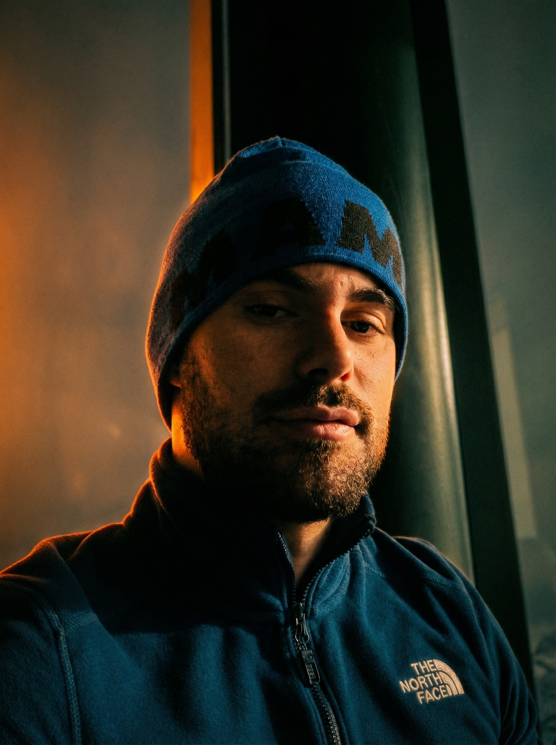 Transform the source image into a high-resolution, moody cinematic film still, while maintaining absolute fidelity to the 100% original subject's facial features, pose, and gender. Keep the person's pose exactly 100% the same as the real image. Perform a hyper-realistic upscale, bringing the subject into extremely sharp focus. Significantly enhance and sharpen skin texture to be incredibly detailed and lifelike, showing visible pores, fine lines, and realistic definition. Apply dramatic, low-key backlighting that casts intense warm orange and deep amber tones as the dominant light source, creating strong rim lighting and a heated glow around the silhouette. The shadows must be crushed deep black, with distinct cool teal and muted dark green undertones visible in the darker background areas. Add a heavy, authentic analog film grain texture over the entire image. Shallow depth of field, high contrast chiaroscuro lighting, atmospheric, emotional, dark cinematic aesthetic. | User Input: N/A