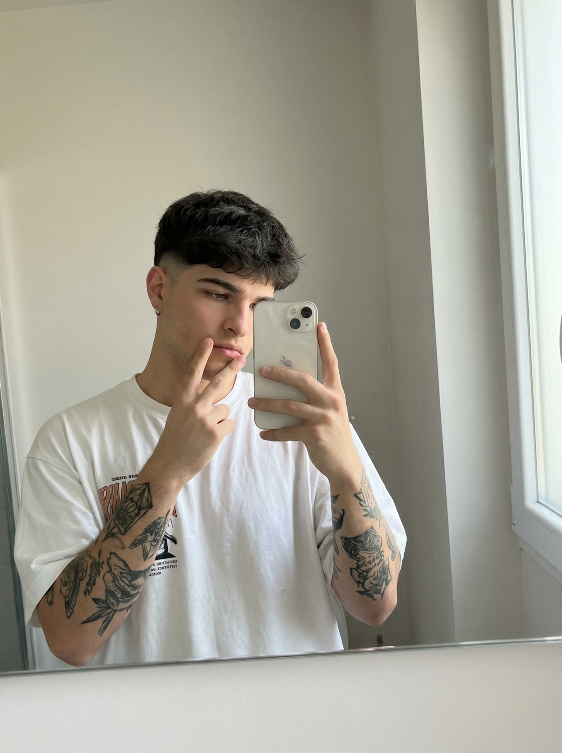 Subject : [blank] The most real and human possible, create an image that looks like a selfie taken with an iPhone in a mirror, captured in portrait orientation with a 2:3 aspect ratio. There should be no clear subject or specific composition — just a casual, unintentional snapshot. The subject holds a light-colored iPhone in one hand and raises the other hand in a relaxed peace sign near the face, standing slightly angled toward a simple bathroom mirror. Keep the exact look from the reference: an oversized white graphic T-shirt, a small hoop earring, and the visible black-ink tattoos covering both forearms, with the tattoo designs clearly preserved as part of the look. The setting is a minimal indoor bathroom with plain light walls, a clean mirror frame, and soft natural-looking ambient light that feels flat and real, like daylight coming through a nearby window. It has slight, natural motion blur from a quick handheld capture. No color grading, no stylization, no cinematic look. The tone must be neutral, not too cool, not too warm. The background must be fully sharp and in focus — no bokeh, no depth-of-field effect, no portrait mode. Framing: off-center, a bit crooked, part of the face sharply captured, with the phone partly blocking the torso in an ordinary mirror-snap composition. Use realistic iPhone camera behavior with subtle indoor softness, authentic skin and fabric texture, and natural mirror reflections. It must not look AI-generated, but rather like it was taken by a real person. [DO NOT CHANGE SUBJECT/S' FACIAL FEATURES] | User Input: N/A