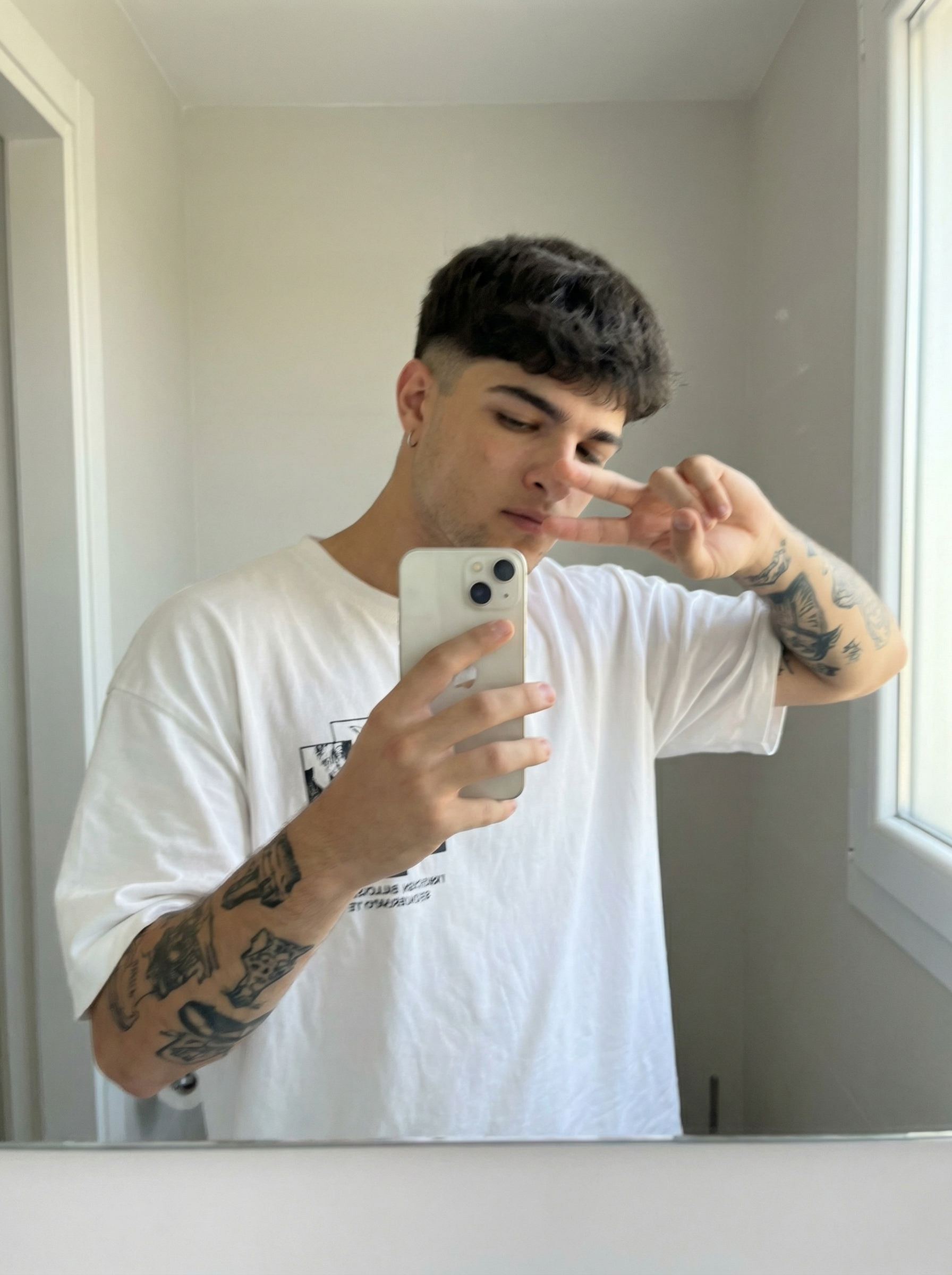 Subject : [blank] The most real and human possible, create an image that looks like a selfie taken with an iPhone in a mirror, captured in portrait orientation with a 2:3 aspect ratio. There should be no clear subject or specific composition — just a casual, unintentional snapshot. The subject holds a light-colored iPhone in one hand and raises the other hand in a relaxed peace sign near the face, standing slightly angled toward a simple bathroom mirror. Keep the exact look from the reference: an oversized white graphic T-shirt, a small hoop earring, and the visible black-ink tattoos covering both forearms, with the tattoo designs clearly preserved as part of the look. The setting is a minimal indoor bathroom with plain light walls, a clean mirror frame, and soft natural-looking ambient light that feels flat and real, like daylight coming through a nearby window. It has slight, natural motion blur from a quick handheld capture. No color grading, no stylization, no cinematic look. The tone must be neutral, not too cool, not too warm. The background must be fully sharp and in focus — no bokeh, no depth-of-field effect, no portrait mode. Framing: off-center, a bit crooked, part of the face sharply captured, with the phone partly blocking the torso in an ordinary mirror-snap composition. Use realistic iPhone camera behavior with subtle indoor softness, authentic skin and fabric texture, and natural mirror reflections. It must not look AI-generated, but rather like it was taken by a real person. [DO NOT CHANGE SUBJECT/S' FACIAL FEATURES] | User Input: N/A