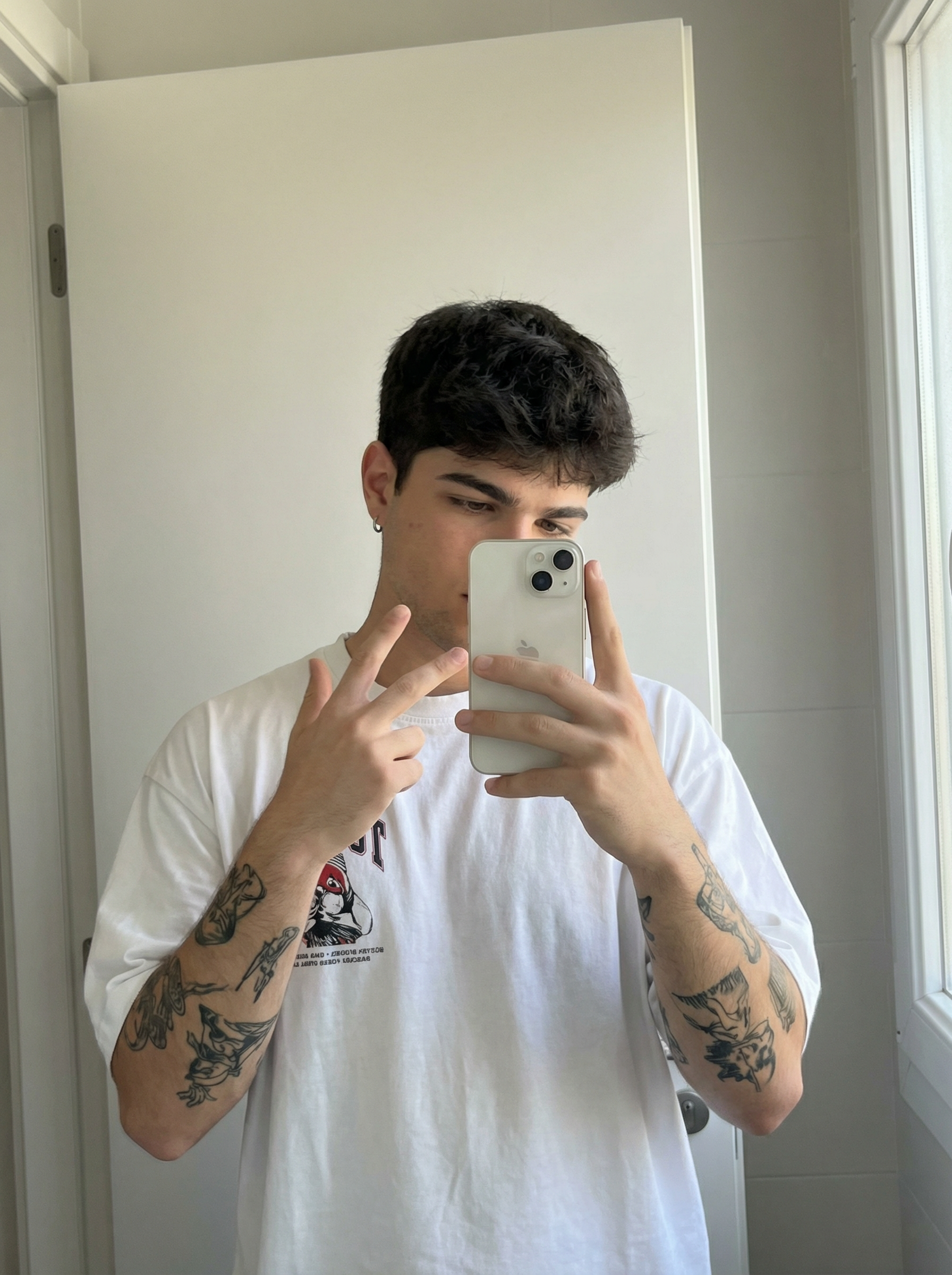 Subject : [blank] The most real and human possible, create an image that looks like a selfie taken with an iPhone in a mirror, captured in portrait orientation with a 2:3 aspect ratio. There should be no clear subject or specific composition — just a casual, unintentional snapshot. The subject holds a light-colored iPhone in one hand and raises the other hand in a relaxed peace sign near the face, standing slightly angled toward a simple bathroom mirror. Keep the exact look from the reference: an oversized white graphic T-shirt, a small hoop earring, and the visible black-ink tattoos covering both forearms, with the tattoo designs clearly preserved as part of the look. The setting is a minimal indoor bathroom with plain light walls, a clean mirror frame, and soft natural-looking ambient light that feels flat and real, like daylight coming through a nearby window. It has slight, natural motion blur from a quick handheld capture. No color grading, no stylization, no cinematic look. The tone must be neutral, not too cool, not too warm. The background must be fully sharp and in focus — no bokeh, no depth-of-field effect, no portrait mode. Framing: off-center, a bit crooked, part of the face sharply captured, with the phone partly blocking the torso in an ordinary mirror-snap composition. Use realistic iPhone camera behavior with subtle indoor softness, authentic skin and fabric texture, and natural mirror reflections. It must not look AI-generated, but rather like it was taken by a real person. [DO NOT CHANGE SUBJECT/S' FACIAL FEATURES] | User Input: N/A