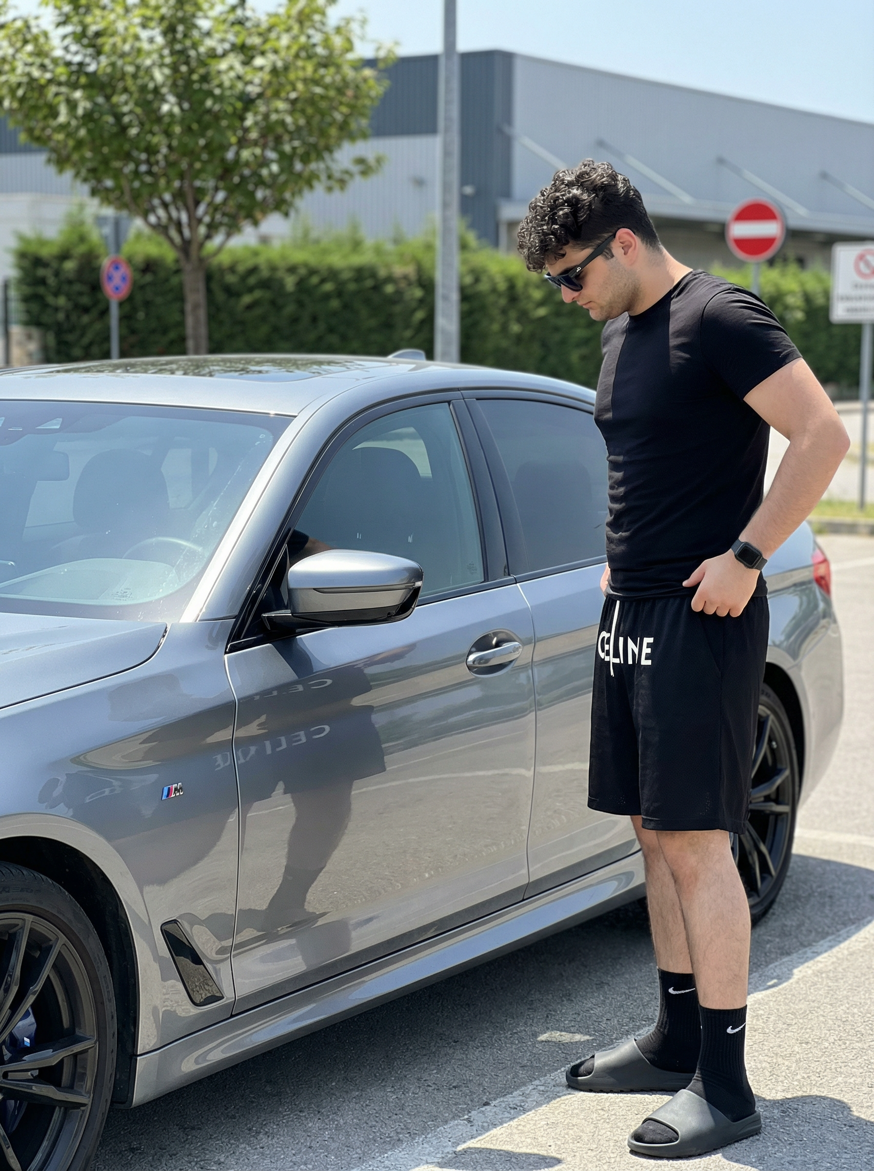 Subject : [blank] Ultra-realistic iPhone 14 Pro Max rear main camera photo at 1x, 24mm equivalent, f/1.78, natural Apple ProRAW look, vertical 9:16, showing the exact same young man standing beside a metallic gray luxury sedan in an outdoor parking area under clear midday daylight, with the framing capturing him nearly full length from head to feet on the right side of the image while the car fills most of the left and center frame, the front wheel cropped off at the far left edge and the rear wheel visible near the right rear of the car; preserve the exact pose with his body turned in clean side profile facing left toward the driver-side door, neck slightly bent forward, chin lowered, gaze directed down toward the car door and window reflection, expression neutral and composed, posture relaxed with a slight forward lean from the shoulders, right hand hooked casually into the waistband or pocket area of his loose black shorts, left arm hanging down naturally with minimal bend, legs straight with a soft bend at the knees and feet planted apart in a casual stance; preserve his exact build, facial structure, skin tone, haircut, and styling, with dark brown hair cut short on the sides in a clean fade and fuller on top with rounded volume, black rectangular sunglasses, a small ear piercing visible, and a fitted black crew-neck T-shirt stretched naturally across the torso with realistic tension and soft fabric cling at the chest, waist, and sleeves; keep the loose knee-length black shorts with white drawstrings hanging at the center and large bold white CELINE lettering across the front, natural folds around the hips and thighs, a black smartwatch on the left wrist, black Nike crew socks with a white swoosh, and dark rubber slide sandals with matte texture; the car should retain the exact visual relationship and proportions, with smooth metallic gray paint, glossy reflections of sky and surroundings on the windows and doors, black multi-spoke wheels, a side vent detail with a small M badge near the front door area, and realistic panel lines, mirror housing, and door handle placement; preserve the surrounding environment with pale textured asphalt, a leafy green tree at upper left, dense green hedge behind the car, a tall dark pole rising near the subject, a red circular no-entry road sign with a white horizontal bar above and behind him, a second partial road sign cut off at the far left edge, and a large industrial or transit-style building in the background with a curved roof and glass-panel façade; lighting must be bright but natural with soft real-world shadow behavior, clean daylight color temperature, restrained smartphone dynamic range, subtle reflections in the car body and windows, crisp focus without artificial sharpening, no portrait mode blur, no cinematic blur, no fake bokeh, no glossy retouching, no editorial stylization, and no CGI feel, keeping the image raw, handheld, believable, and authentically smartphone-real. Maintain the exact appearance, face, and identity of the person from the provided reference image. Do not alter the face. | User Input: N/A