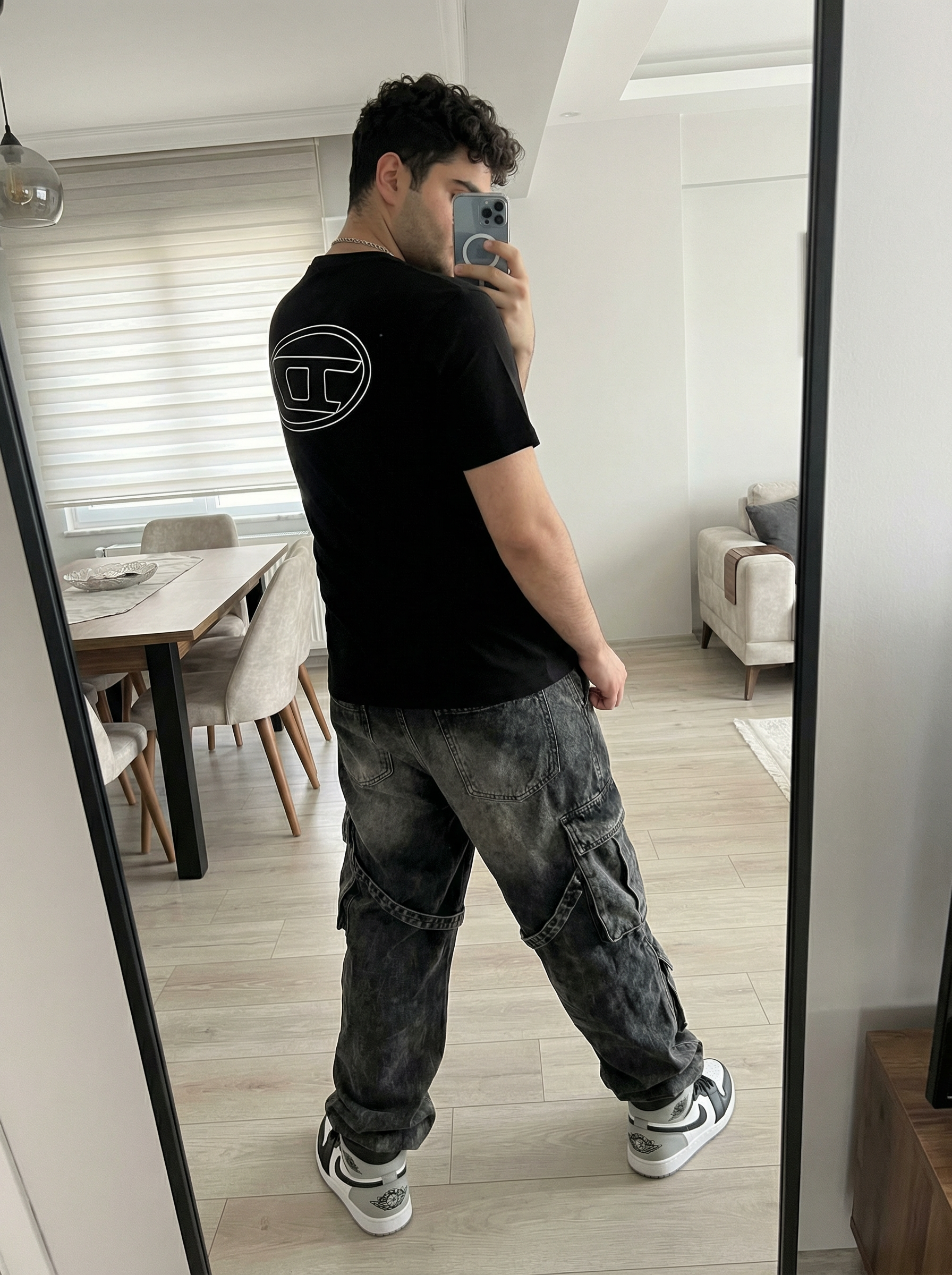 Subject A: [blank]
An ultra-detailed, HD, authentic portrait captured as if taken by an iPhone camera, standing in front of a mirror taking a selfie, with the body angled away from the camera and the back clearly visible, right arm raised holding a smartphone positioned in front of the face, left arm resting naturally down the side, legs positioned in a relaxed, wide stance with weight evenly distributed. The subject is wearing a black short-sleeve t-shirt featuring a large, white outline of the Diesel "D" logo centered on the back, dark grey heavily washed baggy denim cargo pants with large flap pockets on the sides and hanging straps, a silver chain necklace, and black, white, and grey Nike Air Jordan 1 Low sneakers. The scene is reflected in a tall mirror with a thick black curved edge on the right side, the background showing a modern domestic interior with light-colored walls, a dark dining table with black chairs, light wood-plank flooring, a dark rectangular flat-screen TV on the wall, and a window with horizontal blinds letting in natural light. The shot is framed as a full-body mirror reflection from waist level, illuminated by soft ambient indoor lighting with a bright bloom of light emanating from the white smartphone screen being held, casting soft shadows on the clothing and floor. The image is shot on iPhone (Apple ProRAW / RAW-like capture, 12-bit DNG look) as a handheld smartphone capture with a mild wide-angle (~24–28mm equivalent), featuring Smart HDR + Deep Fusion behavior, natural micro-contrast, neutral auto white balance, realistic exposure roll-off, tiny sensor noise, fine grain in shadows, subtle lens distortion, mild vignetting, phone-like depth behavior (not DSLR bokeh), and no beauty filter, no skin smoothing. Avoid AI artifacts: no waxy or plastic textures, no over-sharpening halos, no CGI surfaces, no warped anatomy, no extra fingers, no distorted hands, no fake depth blur. the body pose is an exact 1:1 clone of the reference image, with identical posture, limb positioning, weight distribution, and head tilt, only the subject’s appearance is neutralized while preserving the physical pose perfectly. Do not change Subject's facial features. Subjects must look 1000% identical to uploaded images. no color grading, no stylization, no cinematic look, and the tone must be neutral (not too cool, not too warm). Include subtle iPhone realism cues (mild wide-angle perspective, ~24–28mm equivalent, f/1.9, 1/125s, ISO 125), tiny sensor noise, and natural exposure roll-off; portrait orientation, aspect ratio 4:3, and it must not look AI-generated—just like a real, imperfect iPhone snapshot. | User Input: N/A