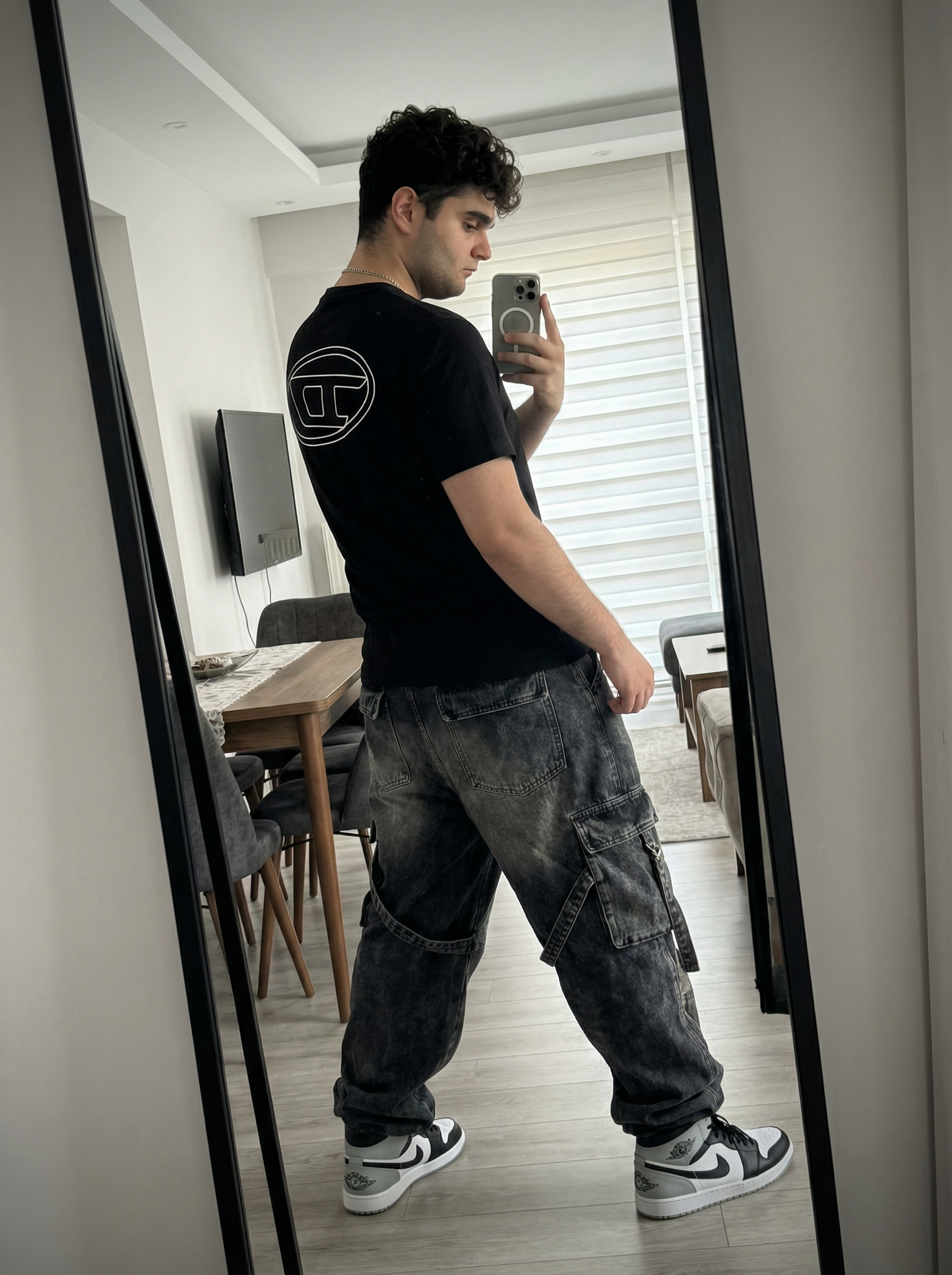 Subject A: [blank]
An ultra-detailed, HD, authentic portrait captured as if taken by an iPhone camera, standing in front of a mirror taking a selfie, with the body angled away from the camera and the back clearly visible, right arm raised holding a smartphone positioned in front of the face, left arm resting naturally down the side, legs positioned in a relaxed, wide stance with weight evenly distributed. The subject is wearing a black short-sleeve t-shirt featuring a large, white outline of the Diesel "D" logo centered on the back, dark grey heavily washed baggy denim cargo pants with large flap pockets on the sides and hanging straps, a silver chain necklace, and black, white, and grey Nike Air Jordan 1 Low sneakers. The scene is reflected in a tall mirror with a thick black curved edge on the right side, the background showing a modern domestic interior with light-colored walls, a dark dining table with black chairs, light wood-plank flooring, a dark rectangular flat-screen TV on the wall, and a window with horizontal blinds letting in natural light. The shot is framed as a full-body mirror reflection from waist level, illuminated by soft ambient indoor lighting with a bright bloom of light emanating from the white smartphone screen being held, casting soft shadows on the clothing and floor. The image is shot on iPhone (Apple ProRAW / RAW-like capture, 12-bit DNG look) as a handheld smartphone capture with a mild wide-angle (~24–28mm equivalent), featuring Smart HDR + Deep Fusion behavior, natural micro-contrast, neutral auto white balance, realistic exposure roll-off, tiny sensor noise, fine grain in shadows, subtle lens distortion, mild vignetting, phone-like depth behavior (not DSLR bokeh), and no beauty filter, no skin smoothing. Avoid AI artifacts: no waxy or plastic textures, no over-sharpening halos, no CGI surfaces, no warped anatomy, no extra fingers, no distorted hands, no fake depth blur. the body pose is an exact 1:1 clone of the reference image, with identical posture, limb positioning, weight distribution, and head tilt, only the subject’s appearance is neutralized while preserving the physical pose perfectly. Do not change Subject's facial features. Subjects must look 1000% identical to uploaded images. no color grading, no stylization, no cinematic look, and the tone must be neutral (not too cool, not too warm). Include subtle iPhone realism cues (mild wide-angle perspective, ~24–28mm equivalent, f/1.9, 1/125s, ISO 125), tiny sensor noise, and natural exposure roll-off; portrait orientation, aspect ratio 4:3, and it must not look AI-generated—just like a real, imperfect iPhone snapshot. | User Input: N/A