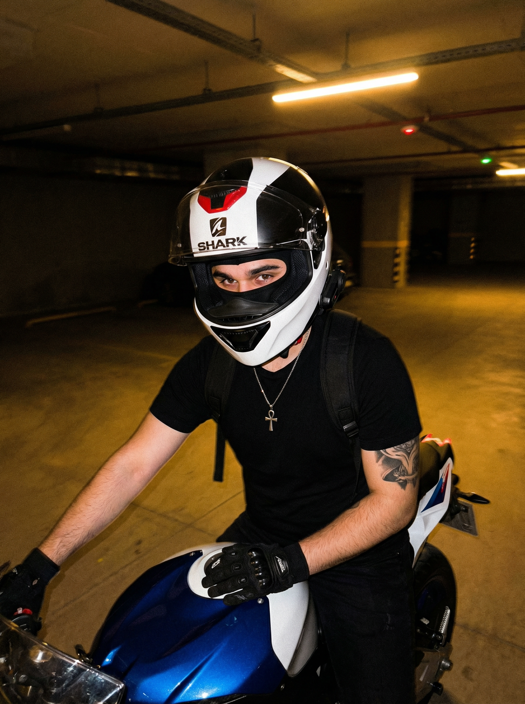 Subject : A person sitting on a motorcycle, wearing a full-face helmet and a black t-shirt.
The most real and human possible, create an image that looks like a casual portrait taken with an iPhone 13 back camera. The raw, amateur, authentic look is paramount. There is no clear subject or specific composition; it's an unintentional, casual snapshot. There is a slight, natural motion blur, and the framing is off-center and a bit crooked, as if taken quickly by a friend. ABSOLUTELY NO AI perfection, no cinematic lighting, no filters or artificial enhancements.
The lighting precisely matches a dimly lit, underground parking garage at night, illuminated by sparse, warm yellowish-orange artificial overhead LED lights. The scene is underexposed, dark, and exhibits noticeable digital noise and grain, typical of low-light iPhone photography. The light source creates harsh shadows and pockets of warmth, especially catching the side of the helmet and the left shoulder/arm. No color grading, no stylization, no cinematic look. STRICTLY FORBIDDEN to add artificial light, fill light, or correct the exposure. The color temperature is distinctly warm and orange.
The entire scene, including the background elements, is fully sharp and in focus — no bokeh, no depth-of-field effect, no portrait mode. The image is a 3:4 portrait aspect ratio, captured with a high-angle, arm-extended framing, looking slightly down onto the subject and the motorcycle.
The person is seated on a white and blue sportbike, leaning slightly forward. Their body is angled gently to the right, away from the camera. The head, encased in a helmet, is turned approximately 45 degrees to the right, looking horizontally across the scene. The left arm is bent at the elbow, gloved hand resting casually on the motorcycle's clear plastic windshield, fingers loosely curled over the edge of the blue tank cover. The right arm is bent, its hand resting out of frame, likely on the person's lap or side.
The person wears a full-face motorcycle helmet, predominantly white with intricate black and red accents and silver details. The brand 'SHARK' is visible in red text on the left side. A dark, slightly raised clear visor is partially open, revealing a black balaclava underneath that covers the entire face. A black communication device with visible buttons and a slender profile is attached to the left side of the helmet. A red chin strap is visible beneath the helmet's chin bar. They are wearing a plain, short-sleeved black cotton t-shirt with natural wrinkles and folds along the torso and under the arm. A black backpack with visible straps is worn over the t-shirt. A silver chain necklace with a small, stylized silver Ankh cross pendant hangs over the t-shirt, resting against the chest. The left hand is covered by a black motorcycle glove with white lettering (appearing to be 'SPARE' or 'SPYDER') and reinforced, textured material, possibly leather or synthetic, with visible stitching. A large, intricate tattoo featuring dark, swirling, organic patterns, possibly depicting a dragon's head or tail near the elbow, covers the left forearm from the elbow downwards. Dark, likely black, pants are visible on the legs, around the knees and thighs, made of a durable, matte fabric.
The setting is a dimly lit, industrial-style concrete parking garage at night. The ground is grey asphalt or worn concrete pavement with faded yellow painted lines, hinting at parking spaces. In the background, robust concrete pillars and the low, flat ceiling of the garage are visible, bathed in the soft, warm, yellowish-orange glow from distant artificial light fixtures, casting long shadows. There are faint architectural details on the concrete walls and possibly blurred lights from what appear to be distant building windows or other garage sections in the far background. The motorcycle is a sleek white and blue sportbike; the tank area is prominent with white plastic fairings and a striking blue central section. The clear plastic windshield is visible, reflecting a faint sheen from the overhead lights. Black handlebars and rearview mirrors are visible in the immediate foreground, partially framing the bottom corners of the shot. The air is still, with no visible weather conditions as it's indoors.
It must not look AI-generated, but rather like it was taken by a real person. DO NOT CHANGE SUBJECT/S' FACIAL FEATURES. | User Input: N/A