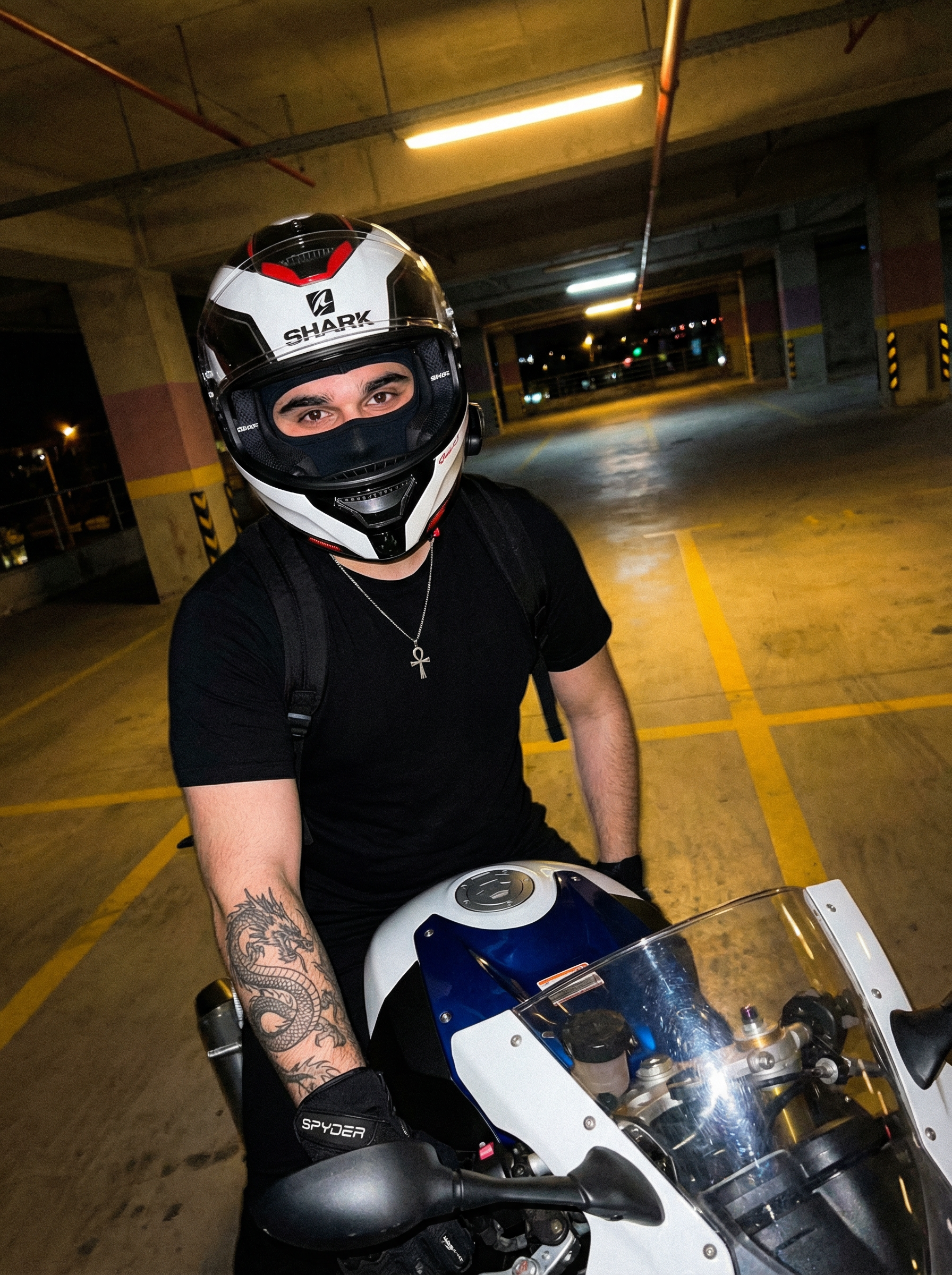 Subject : A person sitting on a motorcycle, wearing a full-face helmet and a black t-shirt.
The most real and human possible, create an image that looks like a casual portrait taken with an iPhone 13 back camera. The raw, amateur, authentic look is paramount. There is no clear subject or specific composition; it's an unintentional, casual snapshot. There is a slight, natural motion blur, and the framing is off-center and a bit crooked, as if taken quickly by a friend. ABSOLUTELY NO AI perfection, no cinematic lighting, no filters or artificial enhancements.
The lighting precisely matches a dimly lit, underground parking garage at night, illuminated by sparse, warm yellowish-orange artificial overhead LED lights. The scene is underexposed, dark, and exhibits noticeable digital noise and grain, typical of low-light iPhone photography. The light source creates harsh shadows and pockets of warmth, especially catching the side of the helmet and the left shoulder/arm. No color grading, no stylization, no cinematic look. STRICTLY FORBIDDEN to add artificial light, fill light, or correct the exposure. The color temperature is distinctly warm and orange.
The entire scene, including the background elements, is fully sharp and in focus — no bokeh, no depth-of-field effect, no portrait mode. The image is a 3:4 portrait aspect ratio, captured with a high-angle, arm-extended framing, looking slightly down onto the subject and the motorcycle.
The person is seated on a white and blue sportbike, leaning slightly forward. Their body is angled gently to the right, away from the camera. The head, encased in a helmet, is turned approximately 45 degrees to the right, looking horizontally across the scene. The left arm is bent at the elbow, gloved hand resting casually on the motorcycle's clear plastic windshield, fingers loosely curled over the edge of the blue tank cover. The right arm is bent, its hand resting out of frame, likely on the person's lap or side.
The person wears a full-face motorcycle helmet, predominantly white with intricate black and red accents and silver details. The brand 'SHARK' is visible in red text on the left side. A dark, slightly raised clear visor is partially open, revealing a black balaclava underneath that covers the entire face. A black communication device with visible buttons and a slender profile is attached to the left side of the helmet. A red chin strap is visible beneath the helmet's chin bar. They are wearing a plain, short-sleeved black cotton t-shirt with natural wrinkles and folds along the torso and under the arm. A black backpack with visible straps is worn over the t-shirt. A silver chain necklace with a small, stylized silver Ankh cross pendant hangs over the t-shirt, resting against the chest. The left hand is covered by a black motorcycle glove with white lettering (appearing to be 'SPARE' or 'SPYDER') and reinforced, textured material, possibly leather or synthetic, with visible stitching. A large, intricate tattoo featuring dark, swirling, organic patterns, possibly depicting a dragon's head or tail near the elbow, covers the left forearm from the elbow downwards. Dark, likely black, pants are visible on the legs, around the knees and thighs, made of a durable, matte fabric.
The setting is a dimly lit, industrial-style concrete parking garage at night. The ground is grey asphalt or worn concrete pavement with faded yellow painted lines, hinting at parking spaces. In the background, robust concrete pillars and the low, flat ceiling of the garage are visible, bathed in the soft, warm, yellowish-orange glow from distant artificial light fixtures, casting long shadows. There are faint architectural details on the concrete walls and possibly blurred lights from what appear to be distant building windows or other garage sections in the far background. The motorcycle is a sleek white and blue sportbike; the tank area is prominent with white plastic fairings and a striking blue central section. The clear plastic windshield is visible, reflecting a faint sheen from the overhead lights. Black handlebars and rearview mirrors are visible in the immediate foreground, partially framing the bottom corners of the shot. The air is still, with no visible weather conditions as it's indoors.
It must not look AI-generated, but rather like it was taken by a real person. DO NOT CHANGE SUBJECT/S' FACIAL FEATURES. | User Input: N/A