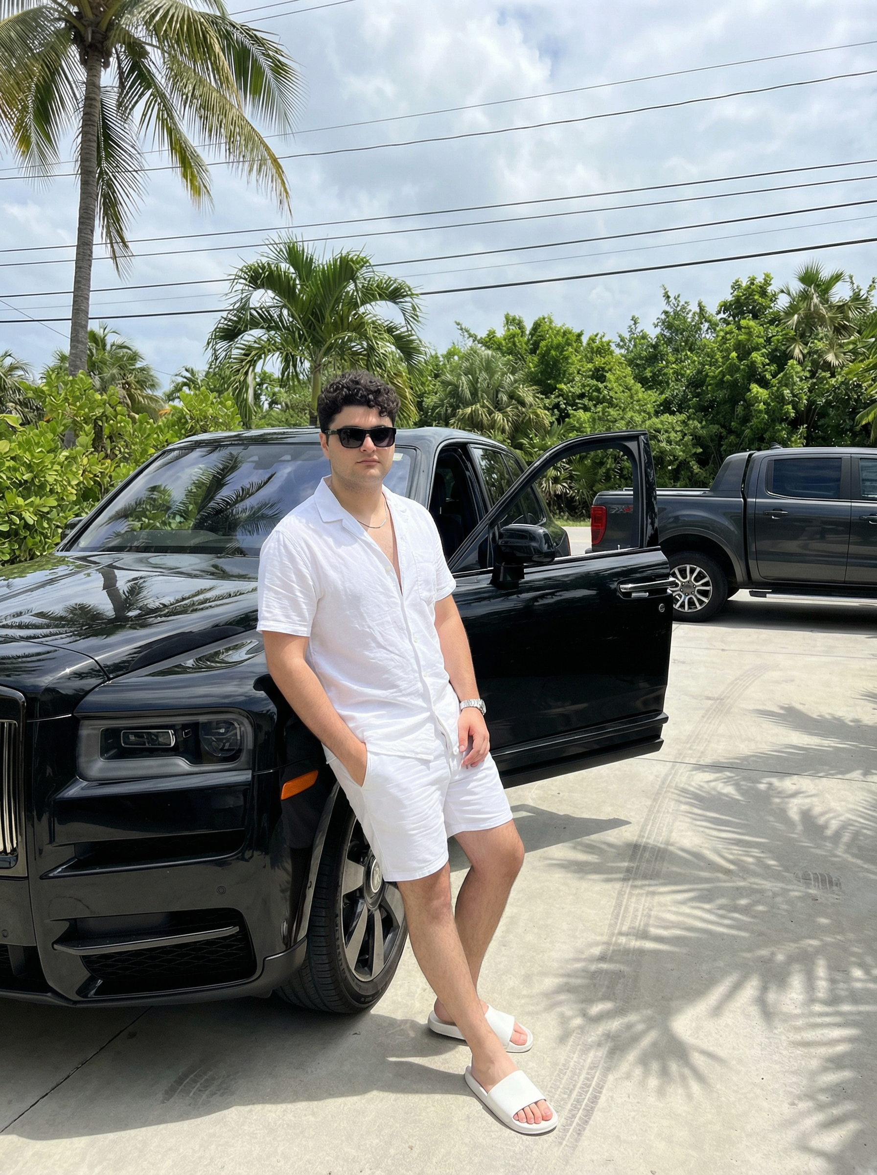 Subject : [blank] The most real and human possible, create an image that looks like a portrait taken by someone else with an iPhone, not a selfie, captured in portrait orientation with a 3:4 aspect ratio. There should be no clear subject or specific composition — just a casual, unintentional snapshot. The subject casually leans against the front corner of a large black luxury SUV with the driver-side door open, one hand resting in a pocket and the other relaxed by the side. Keep the exact look from the reference: a loose short-sleeve white button-up shirt worn open at the collar, matching white shorts, white slide sandals, dark rectangular sunglasses, a thin chain necklace, and a metallic wristwatch. The setting is a tropical outdoor driveway under bright midday sunlight, surrounded by palm trees and dense green foliage with overhead utility wires visible against a partly cloudy sky; another dark pickup truck is parked nearby in the background. The subject lightly interacts with the vehicle by leaning on the front fender while standing on a sunlit concrete surface with subtle tire marks and shadows from the palm leaves overhead. It has slight, natural motion blur from a quick handheld capture. No color grading, no stylization, no cinematic look. The tone must be neutral, not too cool, not too warm. The background must be fully sharp and in focus — no bokeh, no depth-of-field effect, no portrait mode. Framing: off-center, slightly crooked like a friend casually snapped it, capturing part of the SUV grille, the open door, and the surrounding tropical yard. Use realistic iPhone camera behavior with natural daylight exposure around f/2.0, 1/200s shutter, ISO 80–100, subtle palm-leaf shadows across the car surface and ground. It must not look AI-generated, but rather like it was taken by a real person. [DO NOT CHANGE SUBJECT/S' FACIAL FEATURES] | User Input: N/A