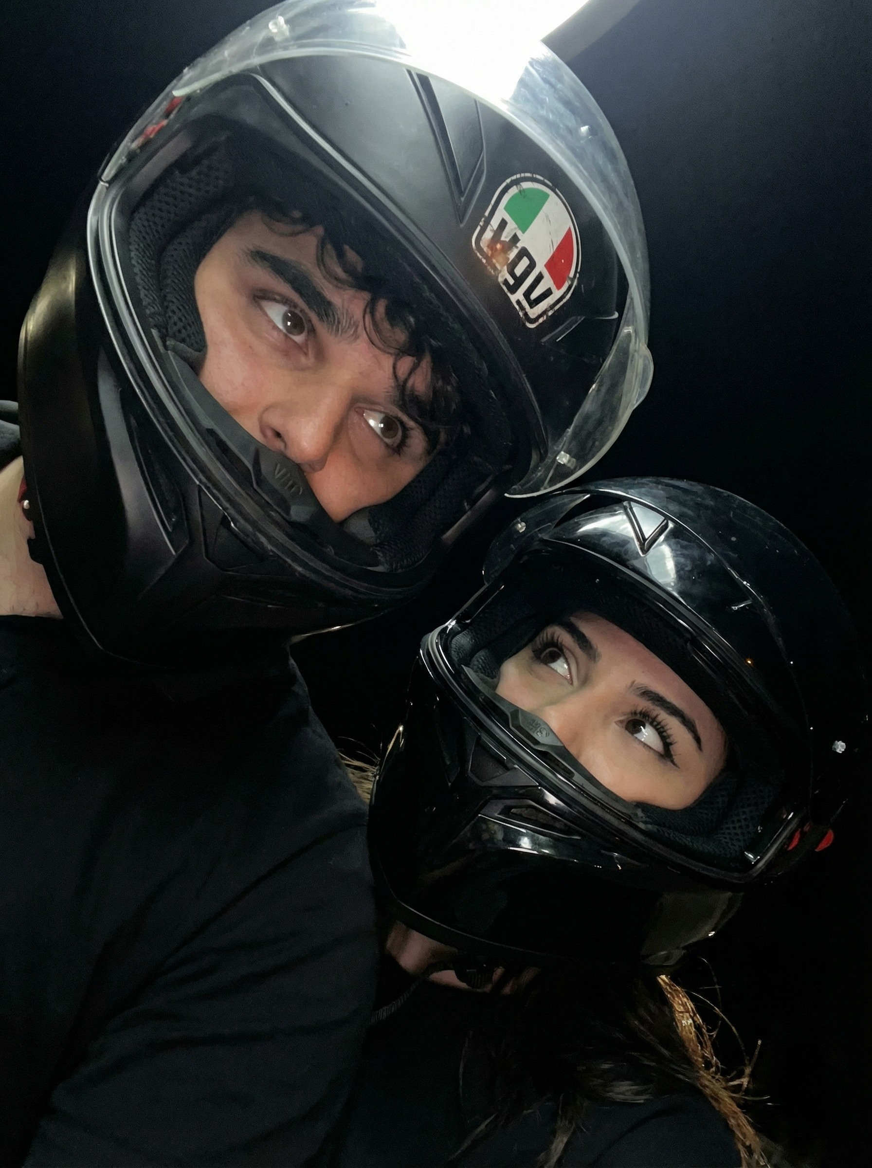 Subject A : [men in the photo]
Subject B : [girl in the photo]
 two individuals wearing motorcycle helmets
The most real and human possible, create an image that looks like a casual portrait taken with an iPhone 13 back camera in portrait orientation (3:4 aspect ratio). The image is a casual, unintentional snapshot, captured with a slightly crooked and off-center high-angle framing, as if taken with an arm slightly extended. There is no clear subject or specific composition, aiming for maximum human imperfection. A slight, natural motion blur is present. No color grading, no stylization, no cinematic look.

The lighting is extremely dark, almost pitch black in the background. The subjects are illuminated by a single, harsh artificial light source from above and slightly to the front, creating high contrast. This results in bright, almost blown-out highlights on the glossy surfaces of the helmets and parts of the visible faces, with deep, underexposed shadows elsewhere. The overall scene appears dark and underexposed, with visible digital noise in the darker areas. No artificial light, no fill light, and no exposure correction is applied.

The foreground subjects (helmets and visible body parts) are fully sharp and in focus. The completely dark background is also implicitly sharp due to its uniform blackness. There is no bokeh, no depth-of-field effect, and no portrait mode.

One individual is positioned at the top-left of the frame, their head and torso angled dynamically, making their black full-face motorcycle helmet appear inverted relative to the frame. The helmet has a matte finish with subtle scratches or imperfections visible. On the left side of this helmet, a distinctive circular "VPG" logo is visible, rendered in red, white, and green with a black outline, showing slight wear. The chin strap is faintly visible. The inner padding of the helmet is black. A portion of the individual's face is visible within the helmet's opening, including the eyes which are looking intensely upwards and slightly to the right. Wavy hair is visible around the forehead and temples, escaping the helmet's inner padding. The left shoulder and upper arm area are visible, covered by a dark (likely black) garment with a visible ribbed or textured pattern, suggesting protective gear.

The second individual is positioned at the bottom-right of the frame, their head tilted slightly upwards and to the left. They are wearing a glossy black full-face motorcycle helmet, its surface highly reflective of the overhead light, showing subtle highlights and reflections. The clear or slightly tinted visor reveals no specific reflections but has a slight sheen. The inner padding of the helmet is black. This individual's eyes are looking upwards and slightly to the left, gazing towards the first individual's general direction. Long, dark eyelashes are prominent. Long hair is visible escaping from the back of the helmet and around the chin strap area. They are wearing a dark (likely black) jacket or top, possibly zipped at the front, with a soft, slightly textured fabric that shows subtle folds and creases. A small sliver of visible skin is present on the neck/chest area just below the helmet's chin bar.

The two helmets are extremely close, almost touching in the center of the frame, suggesting an intimate moment of shared gaze or interaction in the dark environment.

The environment is uniformly pitch black, providing no visible background elements, ground, sky, or weather conditions. There are no reflections other than those on the helmets themselves.

It must not look AI-generated, but rather like it was taken by a real person. DO NOT CHANGE SUBJECT/S' FACIAL FEATURES. | User Input: N/A