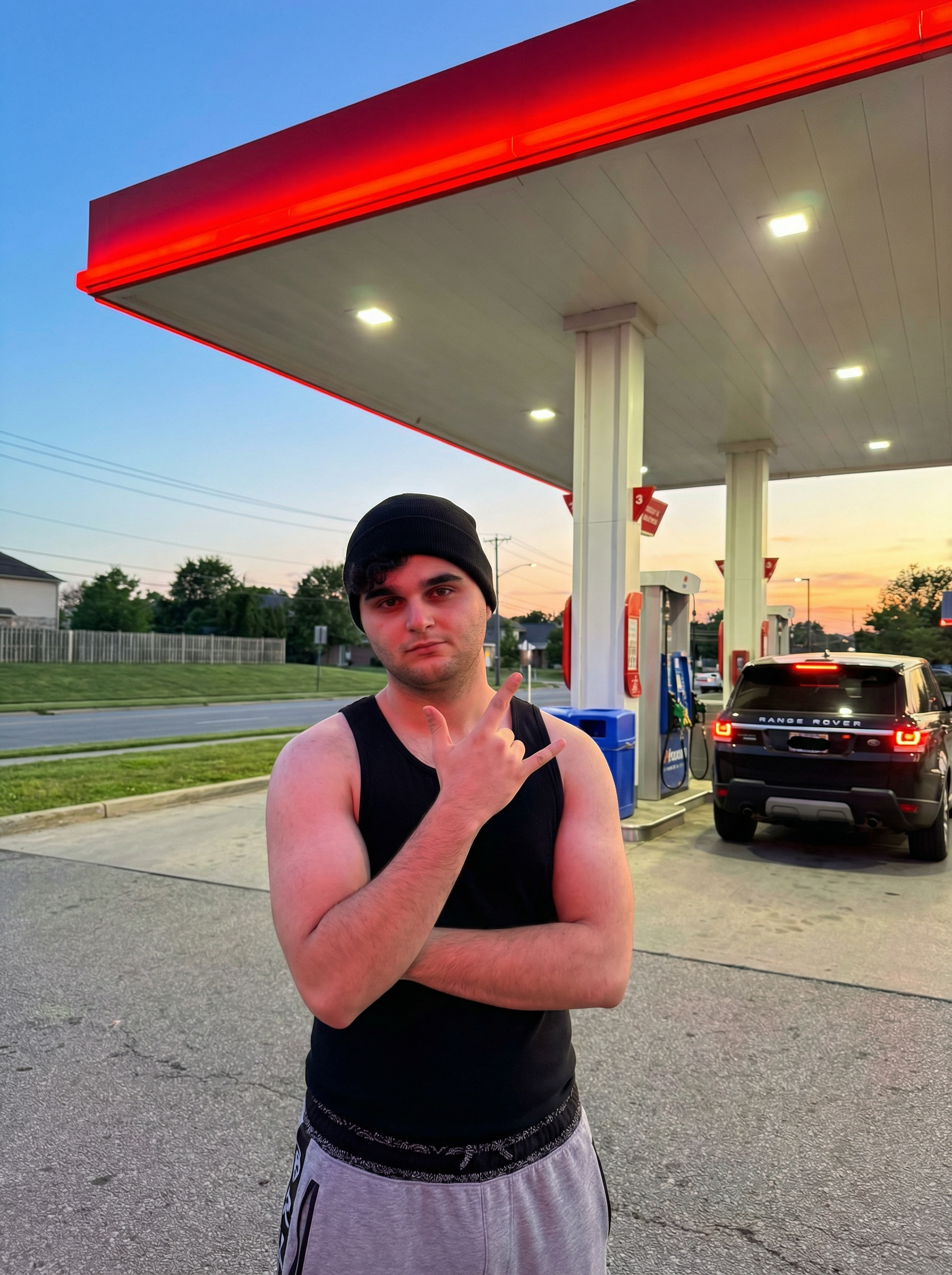 Generate a hyperrealistic image taken with an iPhone 15 Pro Max in HDR mode, 9:16 vertical aspect ratio, natural sunset lighting with blue sky transitioning to orange hues and red glow from the gas station canopy, 8K resolution. The scene is at a gas station with a red illuminated canopy overhead, white support pillars, blue trash bin, gas pumps in the background, a black Range Rover parked nearby with taillights on, grassy area and fence behind, and a suburban road setting at dusk. A person is standing in front of the canopy, arms crossed over the chest with the right hand raised making a hand gesture with index and pinky fingers extended, left arm tucked under, feet shoulder-width apart. The outfit consists of a black beanie on the head, a black sleeveless tank top, gray sweatpants with elastic waistband visible in a patterned black design. The composition places the person in the center foreground, with the gas station elements and car framing the scene, capturing the casual pose in an everyday roadside environment at twilight. | User Input: N/A