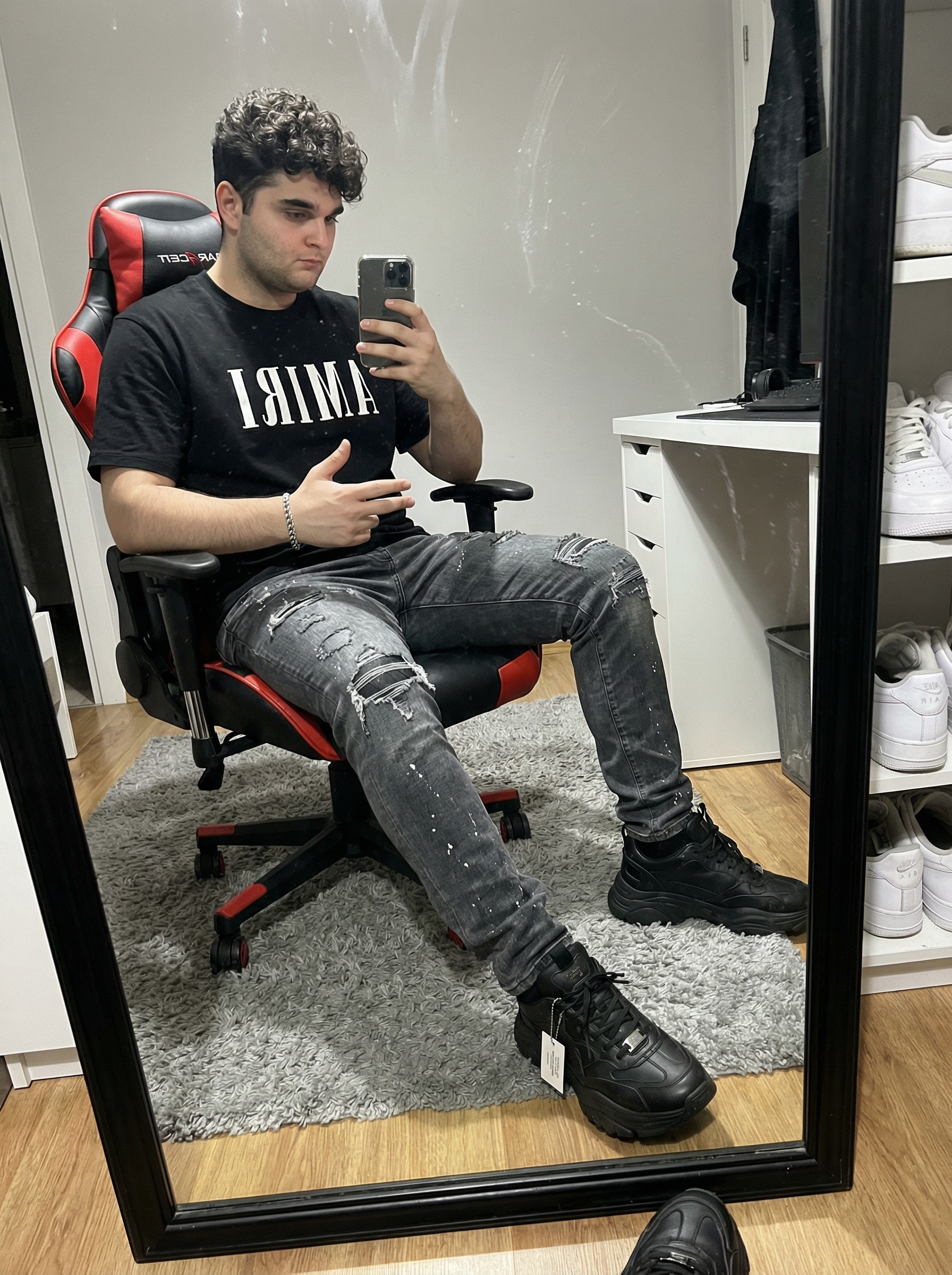 Subject: Person in the image An ultra-detailed, HD, authentic portrait captured as if taken by an iPhone camera, focusing intensely on a mirror selfie. The subject is wearing a heavyweight black cotton t-shirt featuring the "AMIRI" logo in bold white block lettering across the chest. Below, the subject wears heavily distressed charcoal grey denim jeans, characterized by intricate frayed rips at the knees and thighs, accented with subtle white paint splatter textures and faded whiskering. On the feet are chunky black designer sneakers with prominent white contrast stitching and a signature industrial hangtag. A polished silver link bracelet is visible on the wrist. The subject is seated in a red and black ergonomic gaming chair, holding a black smartphone up to capture a reflection in a large, floor-length mirror with a thick black frame. One hand is raised holding the phone, while the other rests on the lap, gesturing toward the camera. The perspective shows the subject's legs extended toward the mirror, with the real feet and lower legs visible in the immediate foreground, creating a sense of depth. The lighting is sharp and direct from an overhead indoor source, creating crisp shadows and a slight sheen on the glossy surface of the mirror. Fine dust particles and faint streaks are visible on the glass surface, adding to the authentic, unedited quality. The background reveals a well-lit room with a grey shag rug on a light wood-grain floor. A white desk with various items and a shelving unit containing white knit sneakers are visible in the reflection, alongside a neutral-toned upholstered headboard. Do not change Subject's facial features. Subjects must look 1000% identical to uploaded images. | User Input: N/A