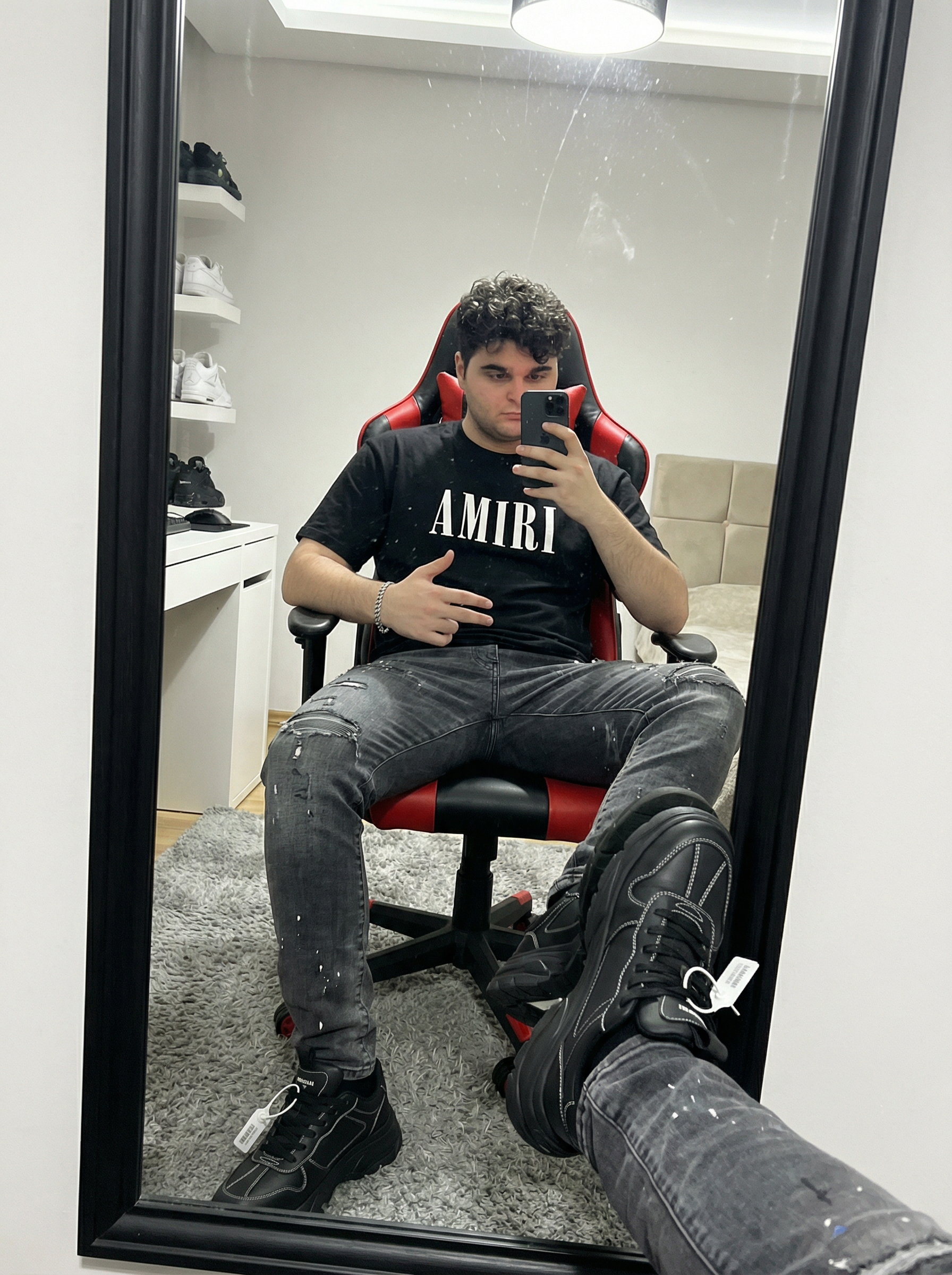Subject: Person in the image An ultra-detailed, HD, authentic portrait captured as if taken by an iPhone camera, focusing intensely on a mirror selfie. The subject is wearing a heavyweight black cotton t-shirt featuring the "AMIRI" logo in bold white block lettering across the chest. Below, the subject wears heavily distressed charcoal grey denim jeans, characterized by intricate frayed rips at the knees and thighs, accented with subtle white paint splatter textures and faded whiskering. On the feet are chunky black designer sneakers with prominent white contrast stitching and a signature industrial hangtag. A polished silver link bracelet is visible on the wrist. The subject is seated in a red and black ergonomic gaming chair, holding a black smartphone up to capture a reflection in a large, floor-length mirror with a thick black frame. One hand is raised holding the phone, while the other rests on the lap, gesturing toward the camera. The perspective shows the subject's legs extended toward the mirror, with the real feet and lower legs visible in the immediate foreground, creating a sense of depth. The lighting is sharp and direct from an overhead indoor source, creating crisp shadows and a slight sheen on the glossy surface of the mirror. Fine dust particles and faint streaks are visible on the glass surface, adding to the authentic, unedited quality. The background reveals a well-lit room with a grey shag rug on a light wood-grain floor. A white desk with various items and a shelving unit containing white knit sneakers are visible in the reflection, alongside a neutral-toned upholstered headboard. Do not change Subject's facial features. Subjects must look 1000% identical to uploaded images. | User Input: N/A