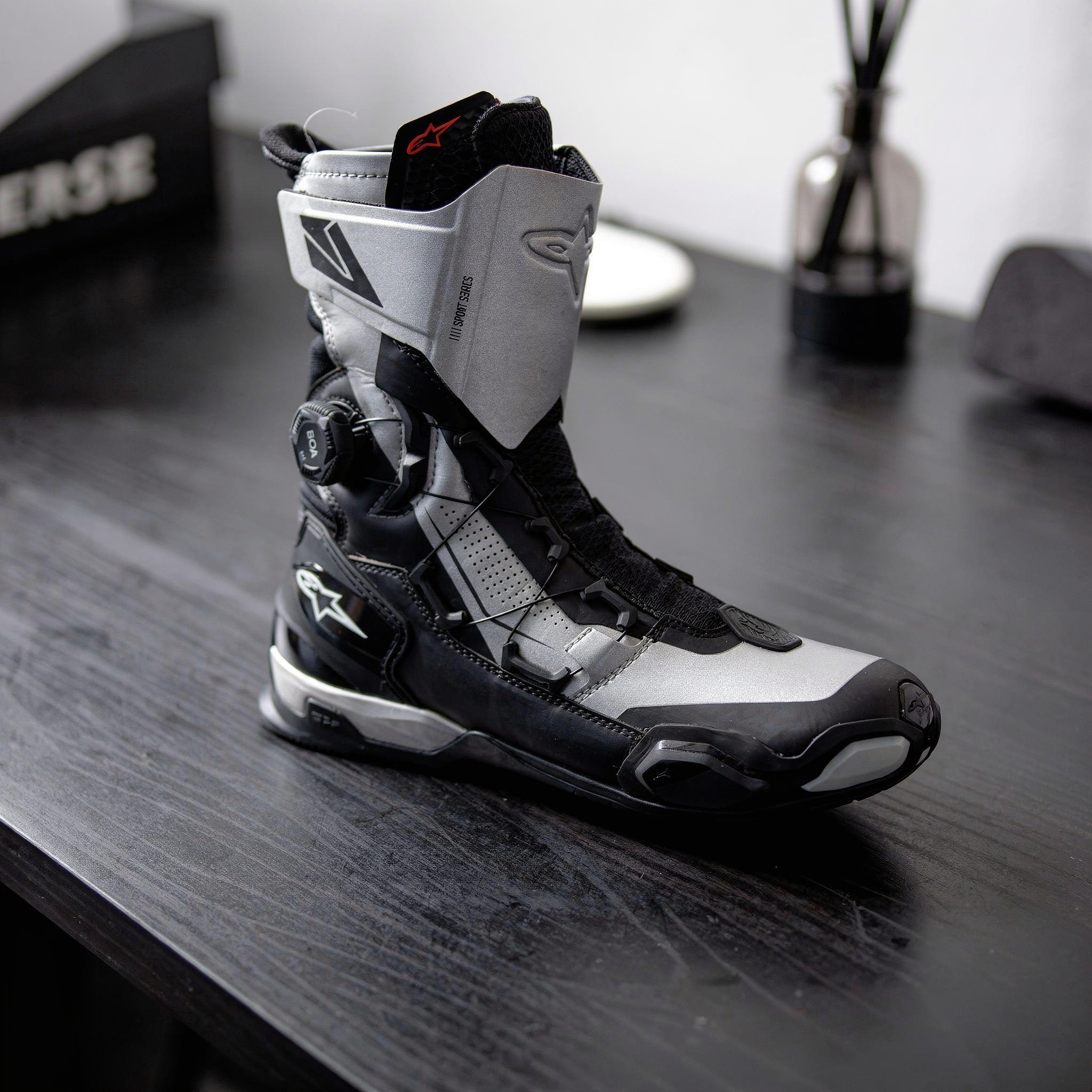 {
  "scene": "A professional product photography setup exactly like the scene in image 1, but with the original sneakers replaced by the motorcycle boots from image 2. The boots are resting on a dark 
