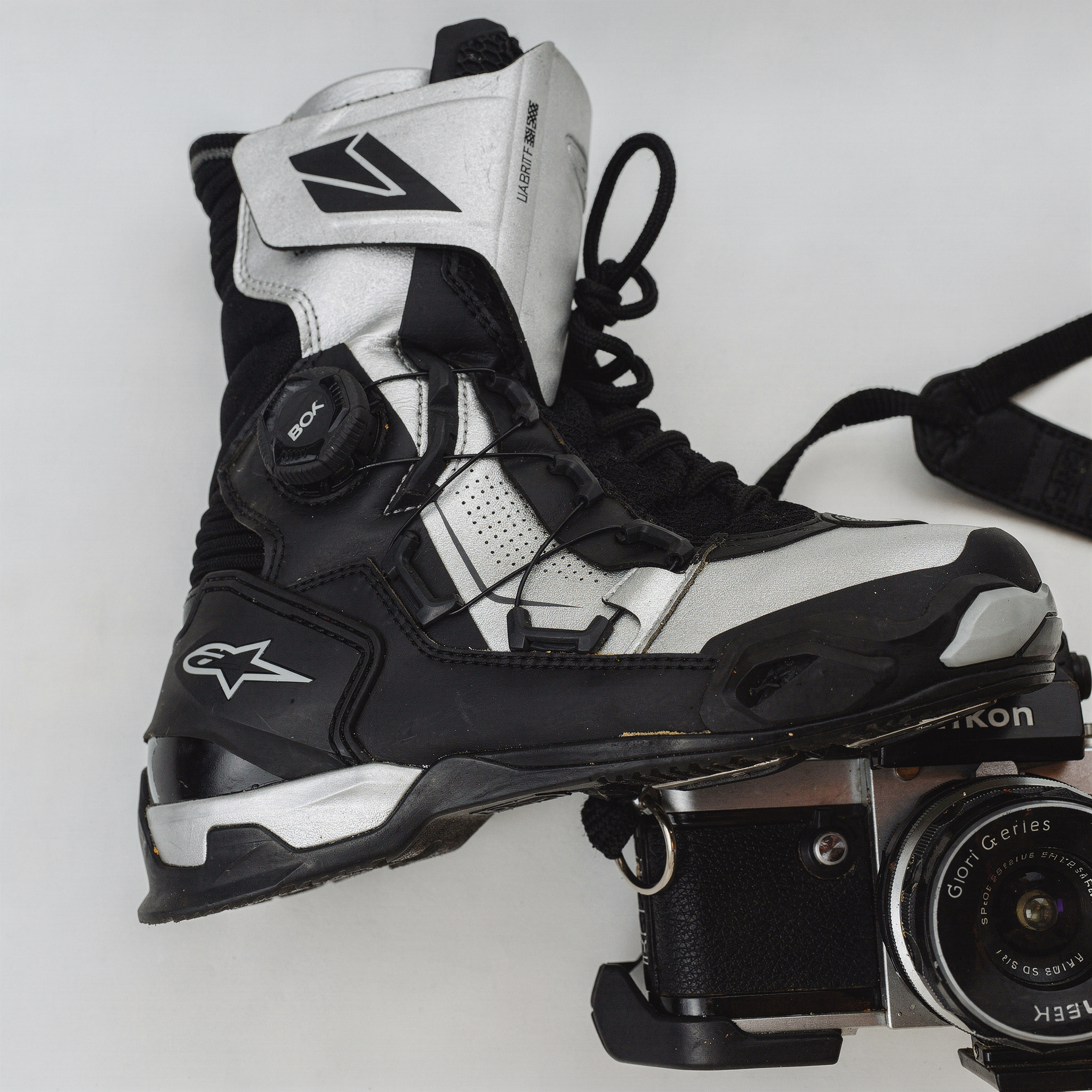 {
  "scene": "A clean, minimalist product photography setup identical to image 1, featuring a stark white surface with a vintage Nikon film camera and its black strap resting on it. The original footw