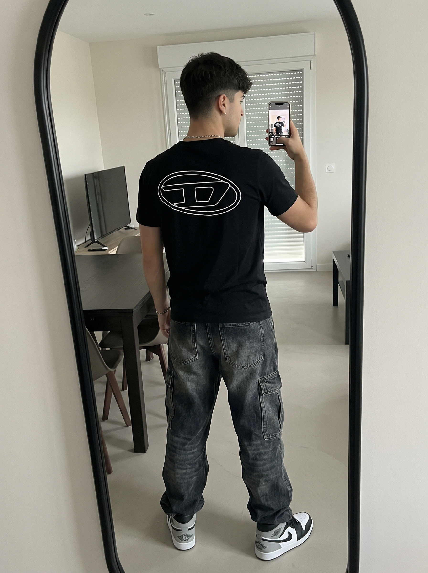 Subject A: [blank]
An ultra-detailed, HD, authentic portrait captured as if taken by an iPhone camera, standing in front of a mirror taking a selfie, with the body angled away from the camera and the back clearly visible, right arm raised holding a smartphone positioned in front of the face, left arm resting naturally down the side, legs positioned in a relaxed, wide stance with weight evenly distributed. The subject is wearing a black short-sleeve t-shirt featuring a large, white outline of the Diesel "D" logo centered on the back, dark grey heavily washed baggy denim cargo pants with large flap pockets on the sides and hanging straps, a silver chain necklace, and black, white, and grey Nike Air Jordan 1 Low sneakers. The scene is reflected in a tall mirror with a thick black curved edge on the right side, the background showing a modern domestic interior with light-colored walls, a dark dining table with black chairs, light wood-plank flooring, a dark rectangular flat-screen TV on the wall, and a window with horizontal blinds letting in natural light. The shot is framed as a full-body mirror reflection from waist level, illuminated by soft ambient indoor lighting with a bright bloom of light emanating from the white smartphone screen being held, casting soft shadows on the clothing and floor. The image is shot on iPhone (Apple ProRAW / RAW-like capture, 12-bit DNG look) as a handheld smartphone capture with a mild wide-angle (~24–28mm equivalent), featuring Smart HDR + Deep Fusion behavior, natural micro-contrast, neutral auto white balance, realistic exposure roll-off, tiny sensor noise, fine grain in shadows, subtle lens distortion, mild vignetting, phone-like depth behavior (not DSLR bokeh), and no beauty filter, no skin smoothing. Avoid AI artifacts: no waxy or plastic textures, no over-sharpening halos, no CGI surfaces, no warped anatomy, no extra fingers, no distorted hands, no fake depth blur. the body pose is an exact 1:1 clone of the reference image, with identical posture, limb positioning, weight distribution, and head tilt, only the subject’s appearance is neutralized while preserving the physical pose perfectly. Do not change Subject's facial features. Subjects must look 1000% identical to uploaded images. no color grading, no stylization, no cinematic look, and the tone must be neutral (not too cool, not too warm). Include subtle iPhone realism cues (mild wide-angle perspective, ~24–28mm equivalent, f/1.9, 1/125s, ISO 125), tiny sensor noise, and natural exposure roll-off; portrait orientation, aspect ratio 4:3, and it must not look AI-generated—just like a real, imperfect iPhone snapshot. | User Input: N/A