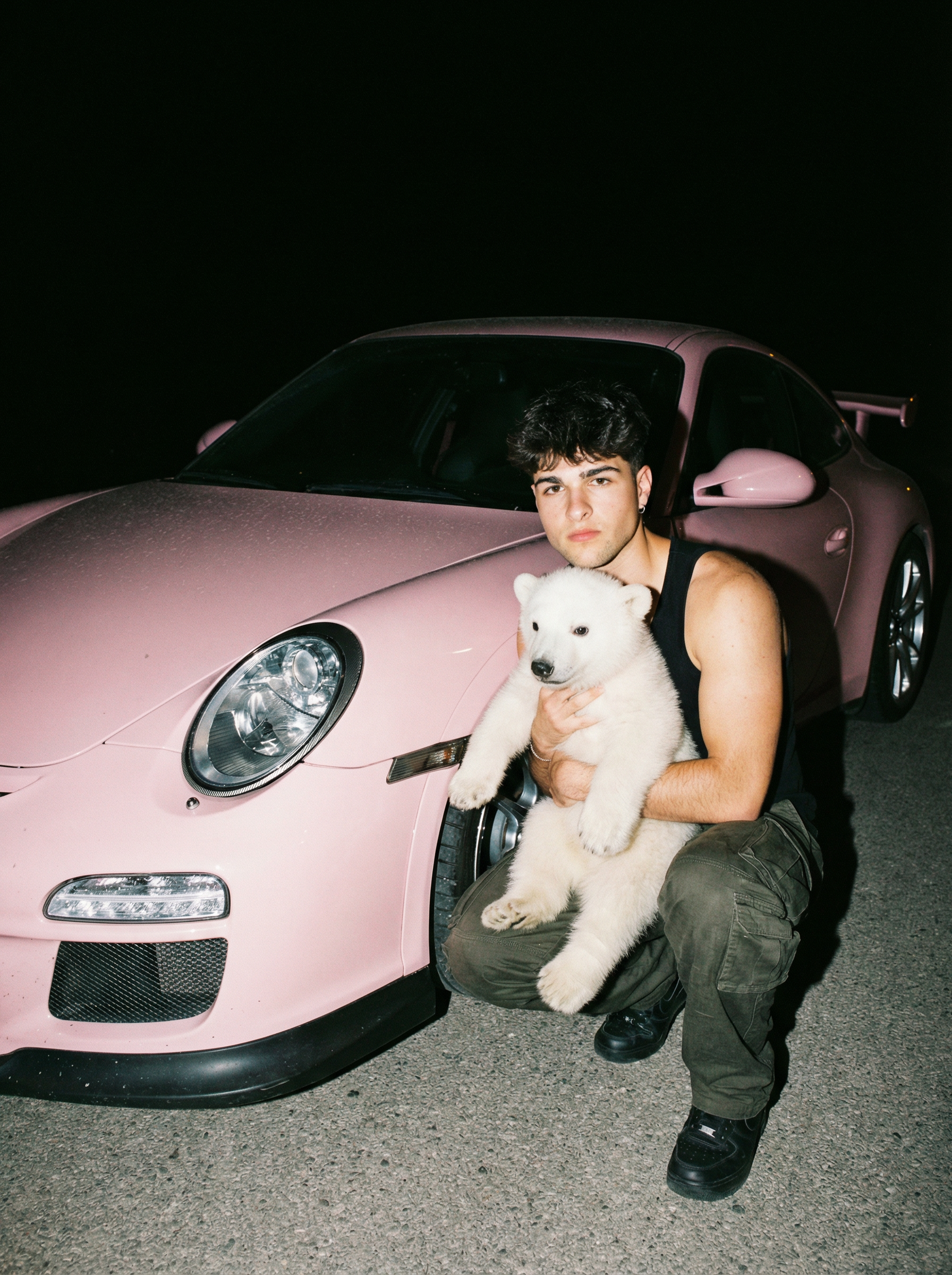 A hyper-realistic nighttime zoom out photograph of a young Māori woman with tattoos, crouching beside a baby pink Porsche GT3 on asphalt, holding a white real polar bear in her arms, wearing military-style cargo pants and a simple top, hair loose and slightly messy, expression neutral and looking directly at the camera without smiling, direct on-camera flash creating sharp highlights on the car's glossy paint and hard shadows behind the subject, the polar bear's fur is catching flash with individual hairs visible, chrome wheel details and Porsche badge reflecting light, dark background with no visible detail beyond the car, cinematic grain, visible skin texture and fabric wrinkles on the cargo pants, tactile detail on the car's paint showing minor dust, documentary fashion photography, intimate and unposed feeling as if shot between moments, shot on 35mm film style, shallow depth of field, raw realism | User Input: N/A