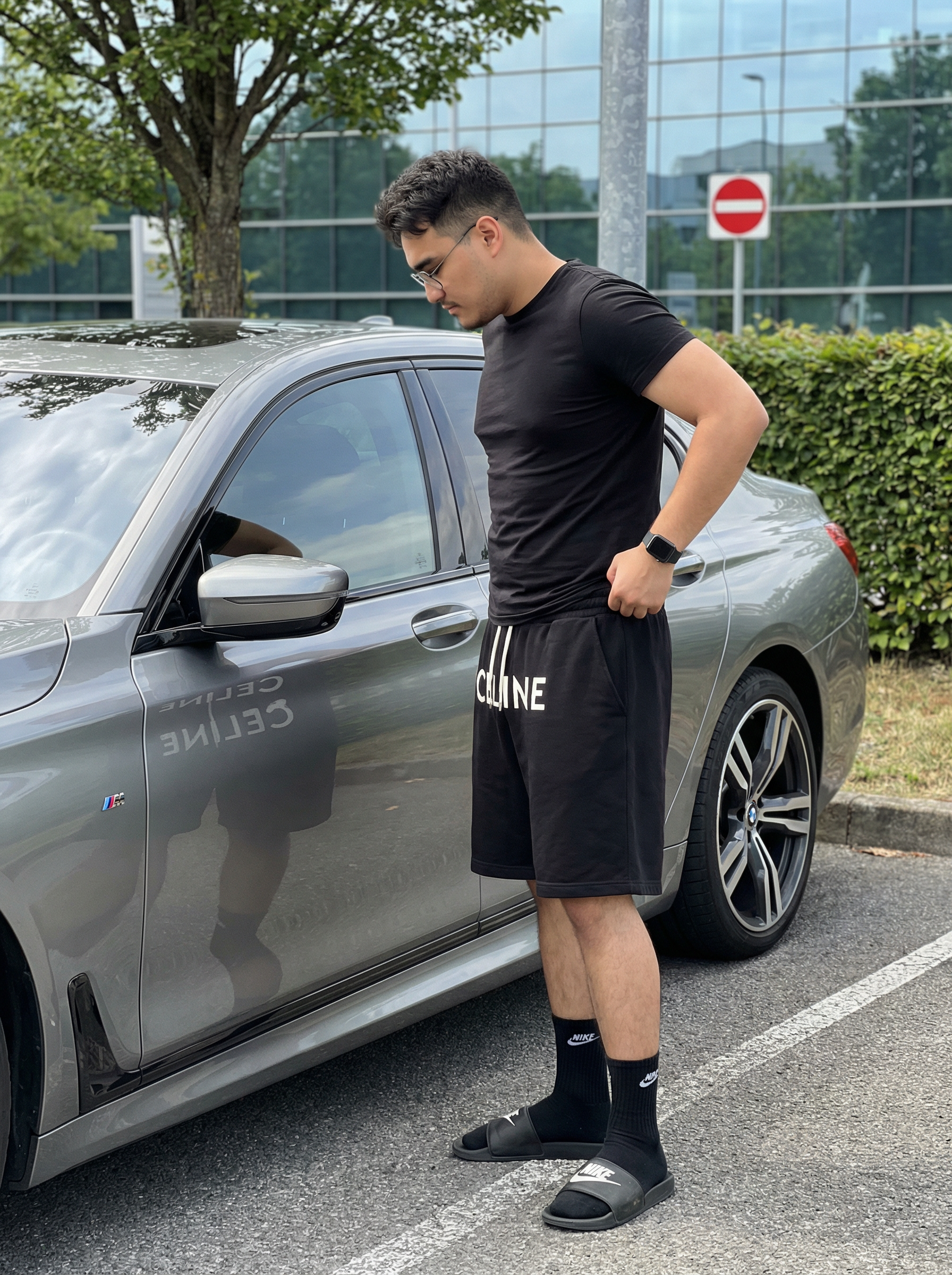 Subject : [blank] Ultra-realistic iPhone 14 Pro Max rear main camera photo at 1x, 24mm equivalent, f/1.78, natural Apple ProRAW look, vertical 9:16, showing the exact same young man standing beside a metallic gray luxury sedan in an outdoor parking area under clear midday daylight, with the framing capturing him nearly full length from head to feet on the right side of the image while the car fills most of the left and center frame, the front wheel cropped off at the far left edge and the rear wheel visible near the right rear of the car; preserve the exact pose with his body turned in clean side profile facing left toward the driver-side door, neck slightly bent forward, chin lowered, gaze directed down toward the car door and window reflection, expression neutral and composed, posture relaxed with a slight forward lean from the shoulders, right hand hooked casually into the waistband or pocket area of his loose black shorts, left arm hanging down naturally with minimal bend, legs straight with a soft bend at the knees and feet planted apart in a casual stance; preserve his exact build, facial structure, skin tone, haircut, and styling, with dark brown hair cut short on the sides in a clean fade and fuller on top with rounded volume, black rectangular sunglasses, a small ear piercing visible, and a fitted black crew-neck T-shirt stretched naturally across the torso with realistic tension and soft fabric cling at the chest, waist, and sleeves; keep the loose knee-length black shorts with white drawstrings hanging at the center and large bold white CELINE lettering across the front, natural folds around the hips and thighs, a black smartwatch on the left wrist, black Nike crew socks with a white swoosh, and dark rubber slide sandals with matte texture; the car should retain the exact visual relationship and proportions, with smooth metallic gray paint, glossy reflections of sky and surroundings on the windows and doors, black multi-spoke wheels, a side vent detail with a small M badge near the front door area, and realistic panel lines, mirror housing, and door handle placement; preserve the surrounding environment with pale textured asphalt, a leafy green tree at upper left, dense green hedge behind the car, a tall dark pole rising near the subject, a red circular no-entry road sign with a white horizontal bar above and behind him, a second partial road sign cut off at the far left edge, and a large industrial or transit-style building in the background with a curved roof and glass-panel façade; lighting must be bright but natural with soft real-world shadow behavior, clean daylight color temperature, restrained smartphone dynamic range, subtle reflections in the car body and windows, crisp focus without artificial sharpening, no portrait mode blur, no cinematic blur, no fake bokeh, no glossy retouching, no editorial stylization, and no CGI feel, keeping the image raw, handheld, believable, and authentically smartphone-real. Maintain the exact appearance, face, and identity of the person from the provided reference image. Do not alter the face. | User Input: N/A