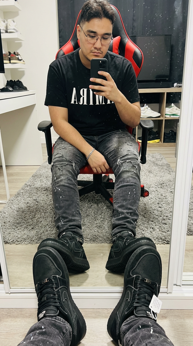 Subject: Person in the image An ultra-detailed, HD, authentic portrait captured as if taken by an iPhone camera, focusing intensely on a mirror selfie. The subject is wearing a heavyweight black cotton t-shirt featuring the "AMIRI" logo in bold white block lettering across the chest. Below, the subject wears heavily distressed charcoal grey denim jeans, characterized by intricate frayed rips at the knees and thighs, accented with subtle white paint splatter textures and faded whiskering. On the feet are chunky black designer sneakers with prominent white contrast stitching and a signature industrial hangtag. A polished silver link bracelet is visible on the wrist. The subject is seated in a red and black ergonomic gaming chair, holding a black smartphone up to capture a reflection in a large, floor-length mirror with a thick black frame. One hand is raised holding the phone, while the other rests on the lap, gesturing toward the camera. The perspective shows the subject's legs extended toward the mirror, with the real feet and lower legs visible in the immediate foreground, creating a sense of depth. The lighting is sharp and direct from an overhead indoor source, creating crisp shadows and a slight sheen on the glossy surface of the mirror. Fine dust particles and faint streaks are visible on the glass surface, adding to the authentic, unedited quality. The background reveals a well-lit room with a grey shag rug on a light wood-grain floor. A white desk with various items and a shelving unit containing white knit sneakers are visible in the reflection, alongside a neutral-toned upholstered headboard. Do not change Subject's facial features. Subjects must look 1000% identical to uploaded images. | User Input: N/A
