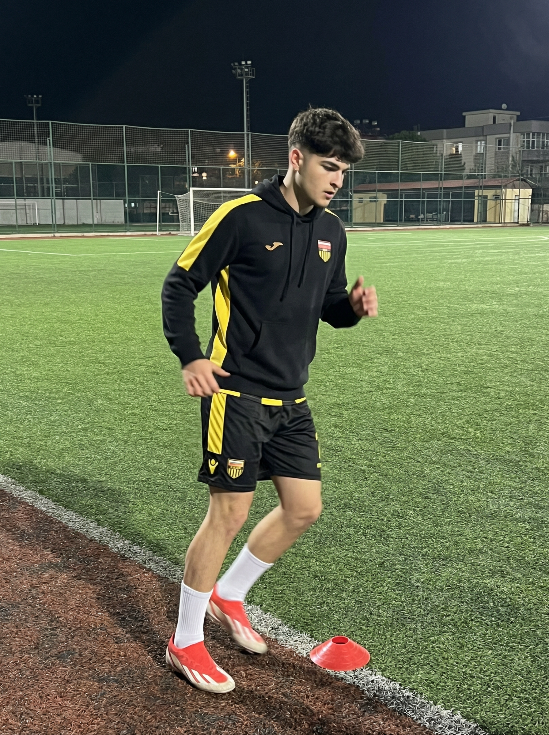 Subject : [blank]
The most real and human possible, create an image that looks like a quick iPhone back-camera portrait taken by a friend from the sideline at night (not a selfie), portrait orientation, aspect ratio 9:16; the subject keeps the exact look from the reference: a black athletic training pullover/hoodie with bold yellow stripe panels running down the sleeves and a small team-style crest patch on the chest, black training shorts with yellow striping and a small yellow text logo near the hem, white mid-calf sports socks, and red-and-white soccer cleats, captured mid-warmup step with arms slightly bent as they move past a small red training cone on an artificial-turf field with painted yard lines and a darker track border. There should be no clear subject or specific composition — just a casual, unintentional snapshot. It has slight, natural motion blur (especially on the moving foot and hands). No color grading, no stylization, no cinematic look. The tone must be neutral, not too cool, not too warm; the scene is lit by bright stadium lights with realistic night exposure (about 26mm equiv, f/1.8–f/2.2, 1/200–1/320s, ISO 800–1600). The background must be fully sharp and in focus — no bokeh, no depth-of-field effect, no portrait mode — showing the field fencing, light poles, and distant structures clearly. Framing: off-center, a bit crooked, part of the face sharply captured; it must not look AI-generated, but rather like it was taken by a real person. [DO NOT CHANGE SUBJECT/S' FACIAL FEATURES] | User Input: N/A