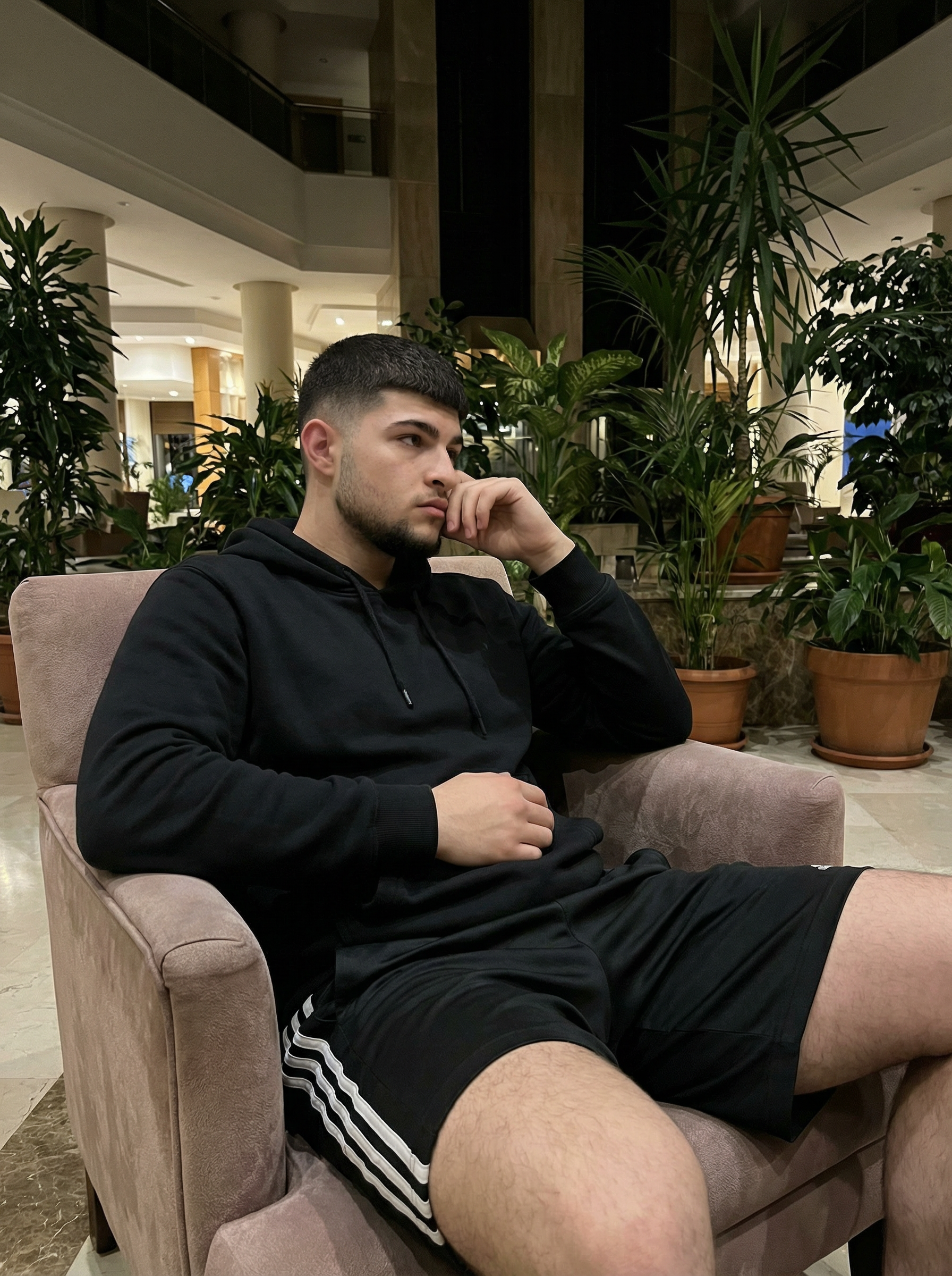 Subject: [Blank]

An ultra-detailed, HD, authentic portrait captured as if taken by an iPhone camera, subject seated in a relaxed, slumped posture, body angled slightly right, head turned sharply left, looking off-camera. Right arm is bent, hand resting on the side of the face near the ear. Left arm is bent, elbow resting on the sofa arm, forearm across the body. Legs are bent and partially visible, implying a relaxed, drawn-up position. Outfit: loose-fitting black hoodie with visible fabric creases, and black athletic shorts featuring two horizontal white stripes on the side seams. Background: lush indoor environment with abundant green foliage and large potted plants, suggesting a hotel lobby or a similar lounge area, with subtle hints of modern architecture and ambient lighting in the distant background. Mid-shot, soft ambient lighting from overhead and background sources, creating a natural, slightly dim appearance with moderate contrast. shot on iPhone (Apple ProRAW / RAW-like capture, 12-bit DNG look), handheld smartphone capture, mild wide-angle (~24–28mm equivalent), Smart HDR + Deep Fusion behavior, natural micro-contrast, neutral auto white balance, realistic exposure roll-off, tiny sensor noise, fine grain in shadows, subtle lens distortion and mild vignetting, phone-like depth behavior (not DSLR bokeh), no beauty filter, no skin smoothing. Avoid AI artifacts: no waxy or plastic textures, no over-sharpening halos, no CGI surfaces, no warped anatomy, no extra fingers, no distorted hands, no fake depth blur. the body pose is an exact 1:1 clone of the reference image, with identical posture, limb positioning, weight distribution, and head tilt, only the subject’s appearance is neutralized while preserving the physical pose perfectly. no color grading, no stylization, no cinematic look, and the tone must be neutral (not too cool, not too warm). Include subtle iPhone realism cues (mild wide-angle perspective, ~24–28mm equivalent, f/1.9, 1/125s, ISO 125), tiny sensor noise, and natural exposure roll-off; portrait orientation, aspect ratio 4:3, and it must not look AI-generated—just like a real, imperfect iPhone snapshot. | User Input: N/A