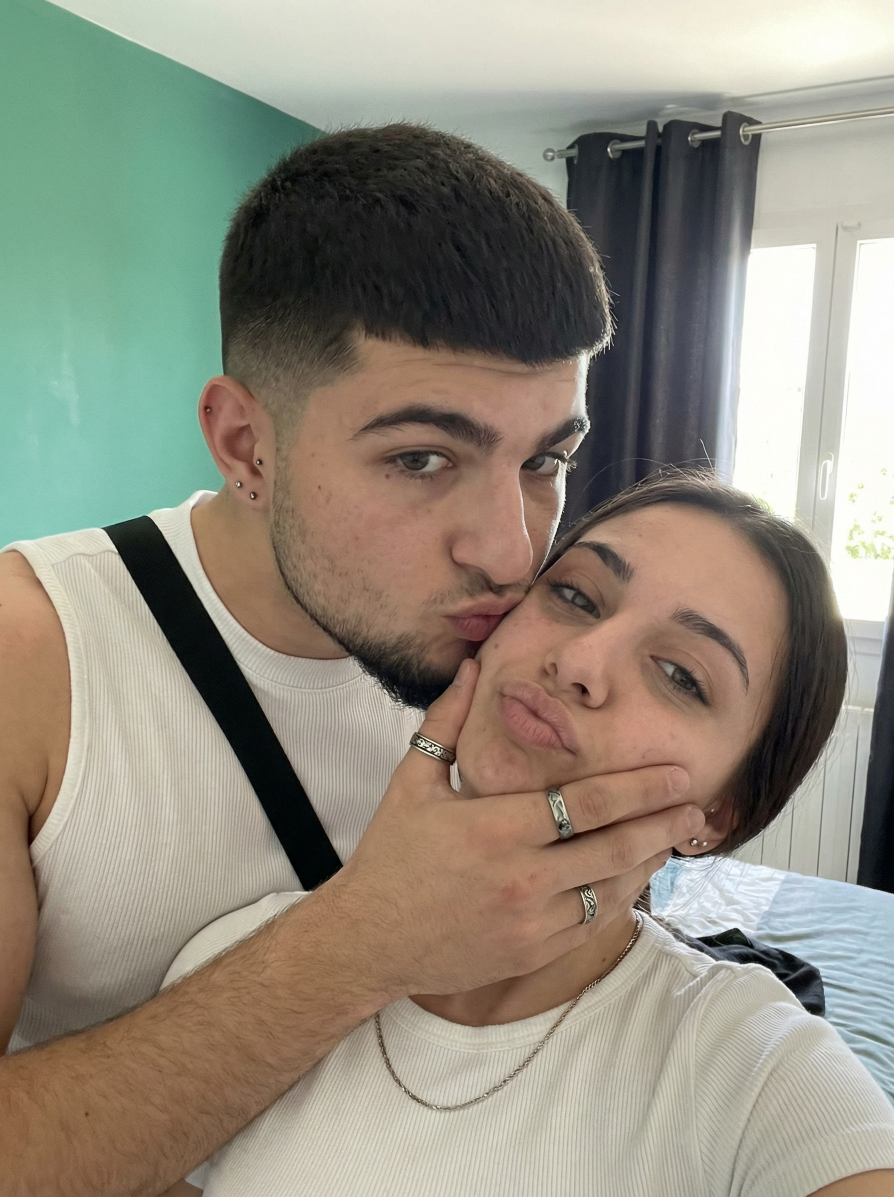 Subject A: [blank]
Subject B: [blank]

The most real and human possible, create an image that looks like a close-up couple selfie taken with an iPhone front camera at arm’s length in portrait orientation, aspect ratio 3:4; Subject A is slightly above/behind, leaning down with lips puckered while wrapping an arm around Subject B and gently holding Subject B’s cheek, with rings on the fingers and small ear jewelry visible, keeping the exact outfit/accessories: a ribbed white sleeveless top with a black underlayer/strap visible. Subject B is lower in frame with a relaxed pout, keeping the exact outfit/accessories: a light-colored (white) ribbed top and a thin chain necklace. The setting is an indoor bedroom/living space in daylight with a teal/green wall and dark curtains near a bright window, soft natural light spilling in and creating mild highlights on skin and fabric texture; realistic iPhone selfie exposure (about 24–28mm equiv, f/1.8–f/2.2, 1/120s, ISO 80–250). There should be no clear subject or specific composition — just a casual, unintentional snapshot. It has slight, natural motion blur. No color grading, no stylization, no cinematic look. The tone must be neutral, not too cool, not too warm; The background must be fully sharp and in focus — no bokeh, no depth-of-field effect, no portrait mode. Framing: off-center, a bit crooked, part of the face sharply captured; all images aspect ratio should be 3:4. It must not look AI-generated, but rather like it was taken by a real person. [DO NOT CHANGE SUBJECT/S' FACIAL FEATURES] | User Input: N/A