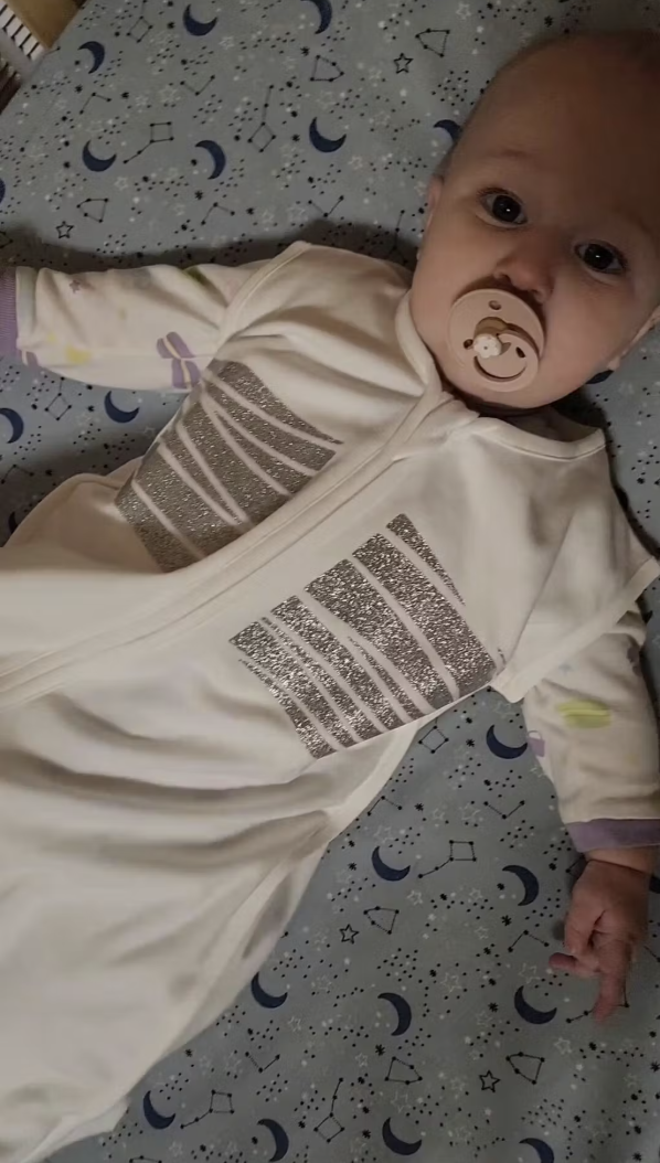 A real customer photo: newborn in the Sleep of Mind sleep sack on a moon-print sheet, silver reflective stripes catching the soft light.