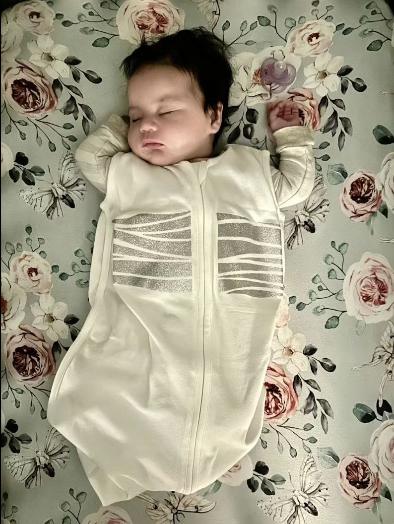 A second real customer photo: the same sleep sack on a different baby, this time on a floral sheet, silver stripes clearly visible across the chest.