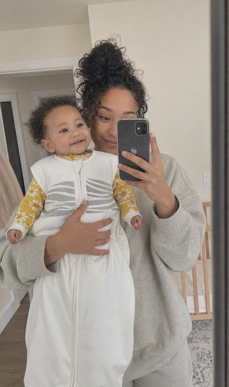 A real Sleep of Mind customer at home with her baby, wearing the cream sleep sack with reflective stripes, mum holding the phone for a mirror selfie.