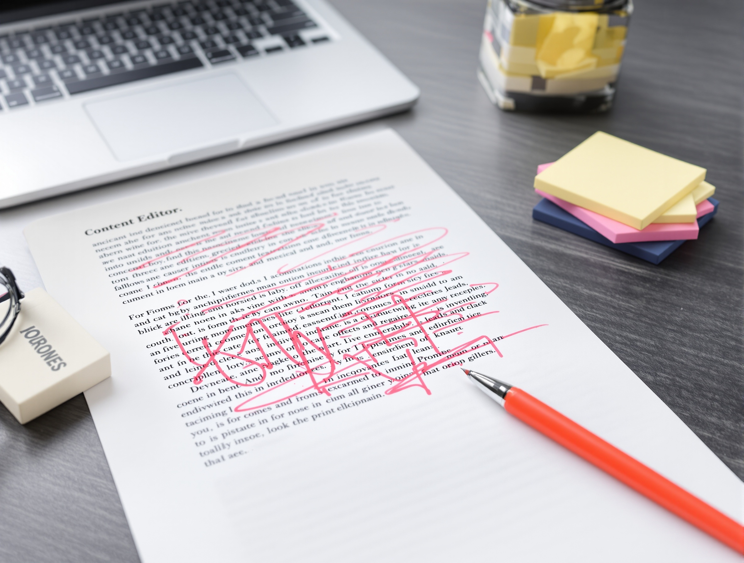 An edited printed draft marked up in red pen next to a laptop on a desk