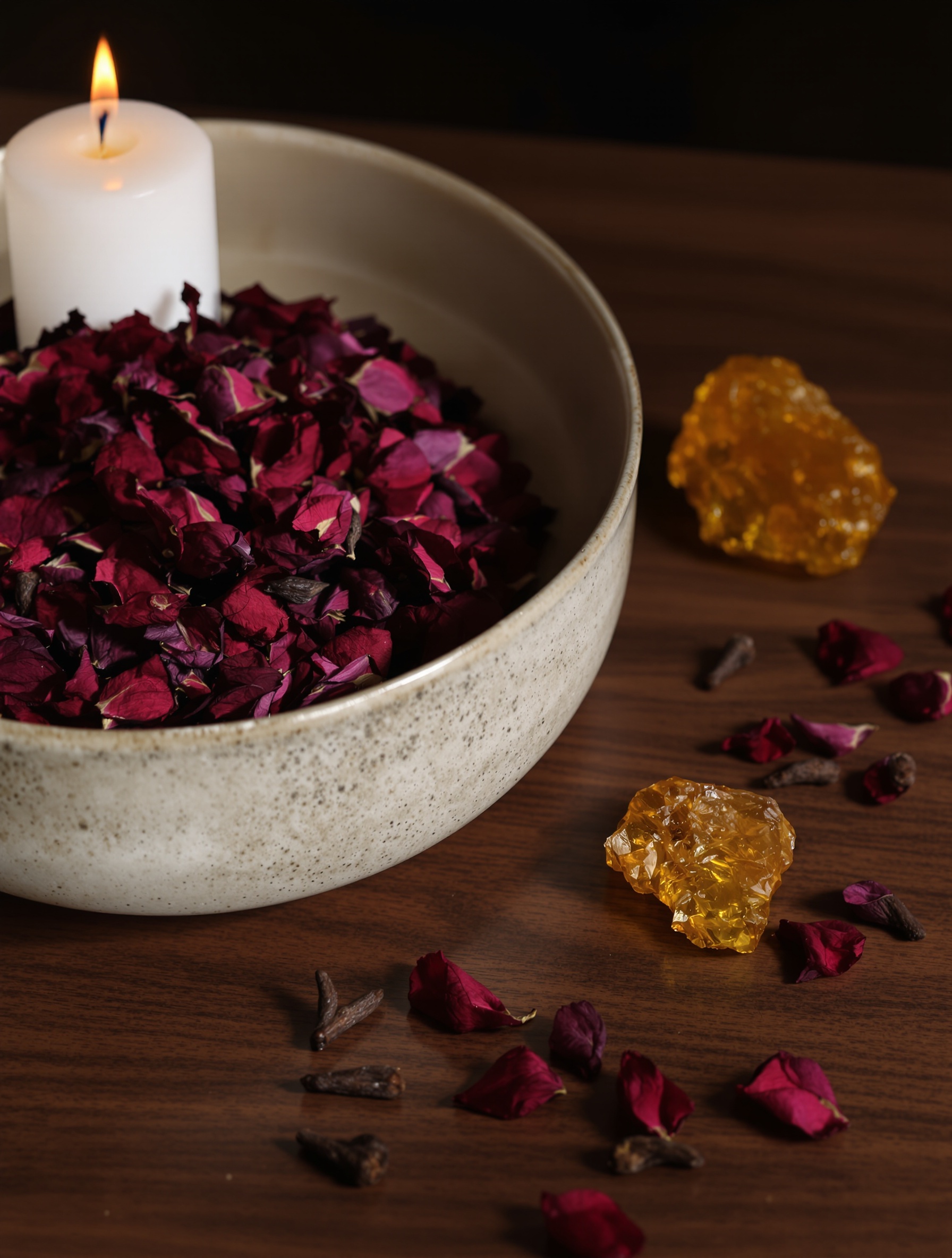 Dried dark rose petals and raw amber resin in a ceramic bowl on mahogany, lit by candlelight, reflecting the dense material heart of Portrait of a Lady.