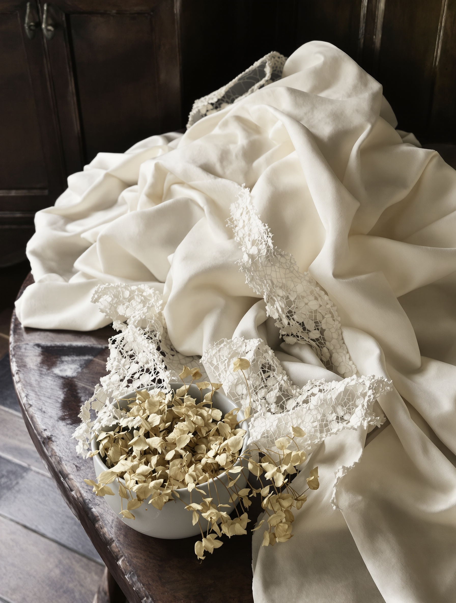 Bolts of aged silk and dried florals on a Victorian receiving-room table, the textile world that surrounds Millésime 1849.