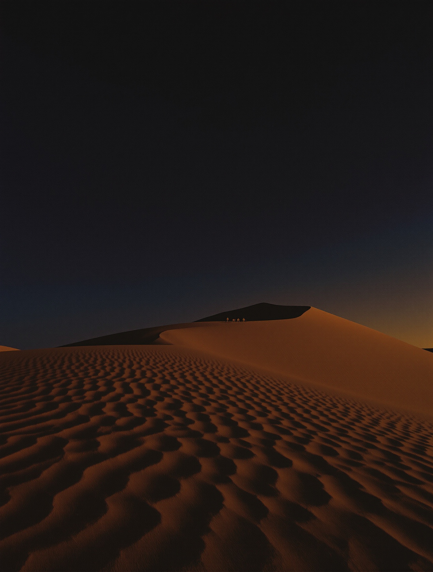 A film-photographed Moroccan desert at deep dusk with a natural indigo sky, windswept sand ripples in amber foreground light, and a tiny camel caravan silhouette on the far horizon.