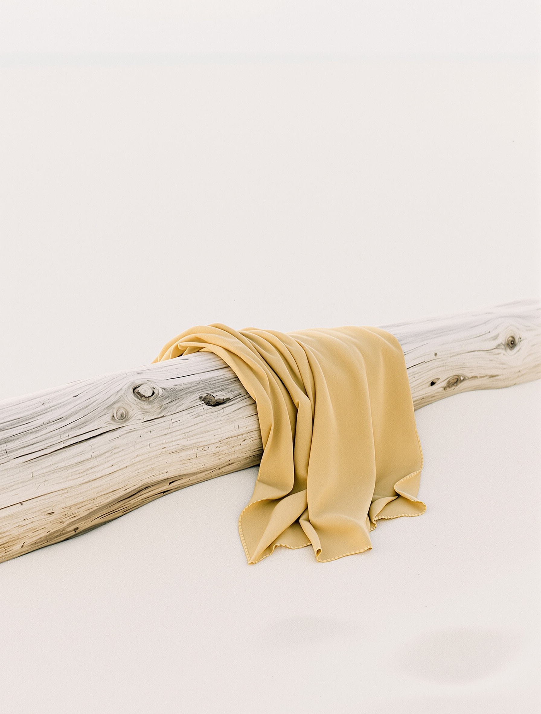 A sun-warmed amber sarong draped over bleached driftwood on a pale empty beach, fabric and sand dissolving into overexposed afternoon light.