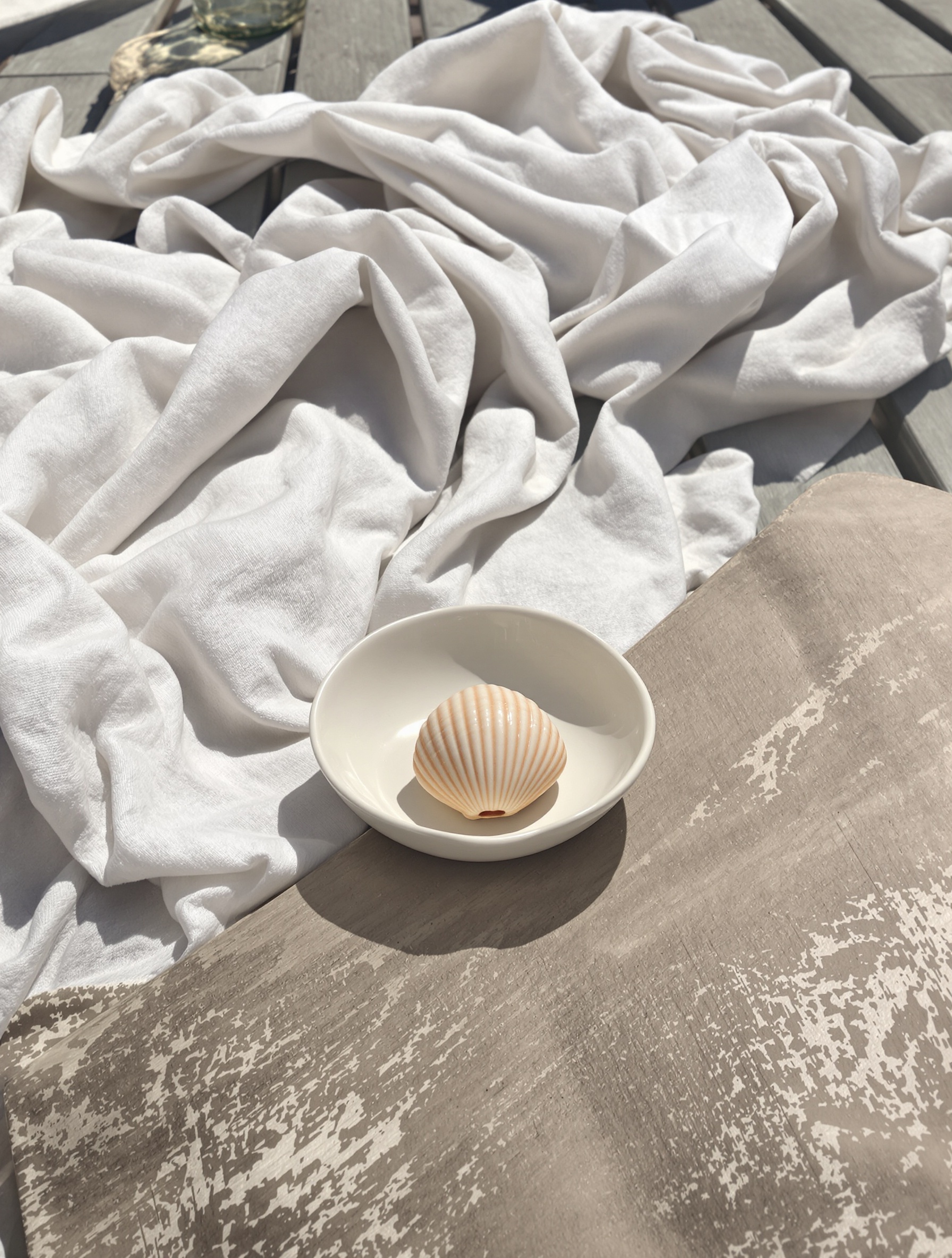 An off-centre ceramic dish with a small ridged shell rests on a sun-bleached teak bench beside rumpled linen, bathed in overexposed afternoon light.