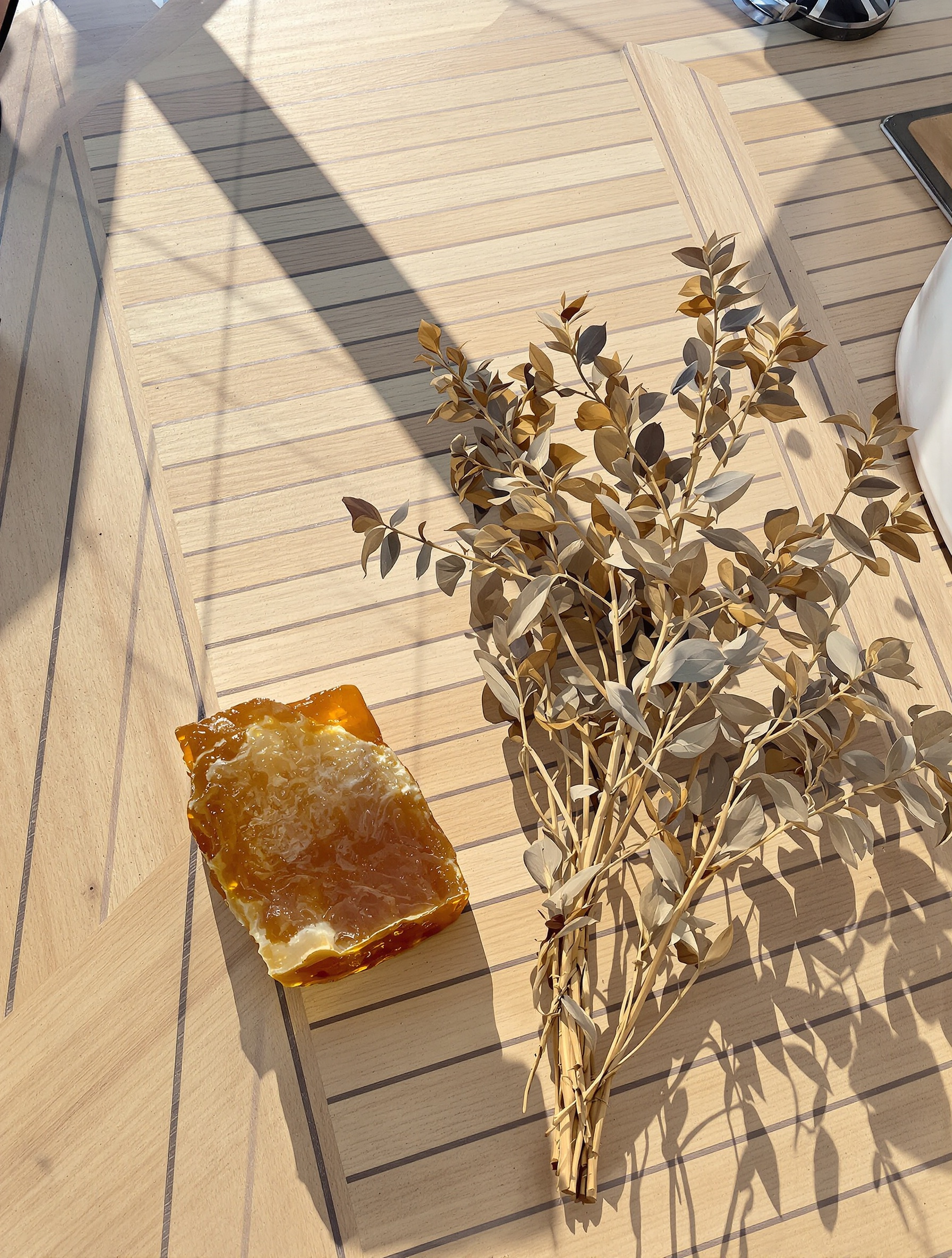 Fragrance ingredients scattered on sun-bleached teak yacht decking under bright coastal daylight, with a hazy sea horizon softly visible beyond.