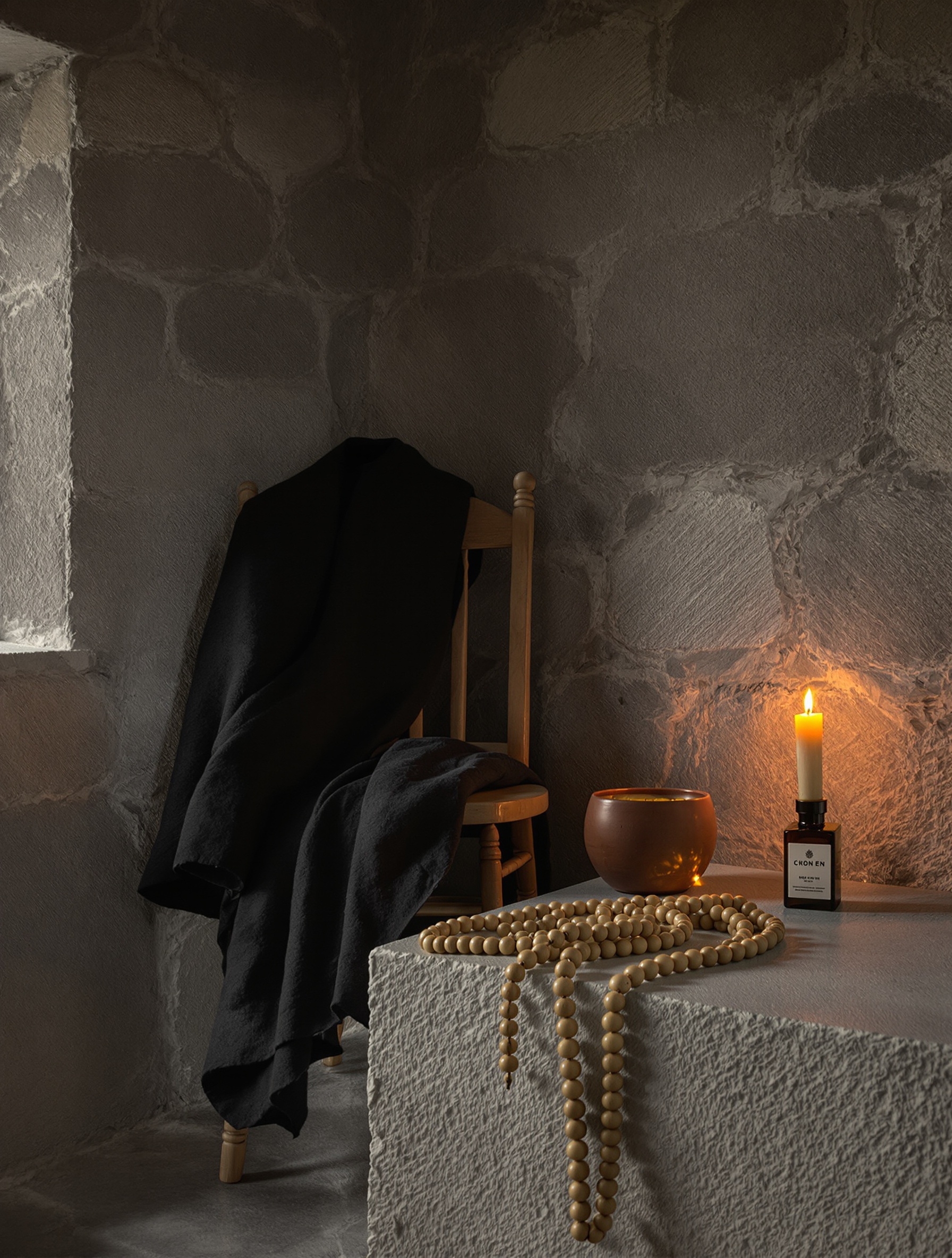 A candlelit stone room holds a dark wool coat draped over a weathered wooden chair beside a clay bowl of amber resin and coiled wooden prayer beads on a rough stone ledge.