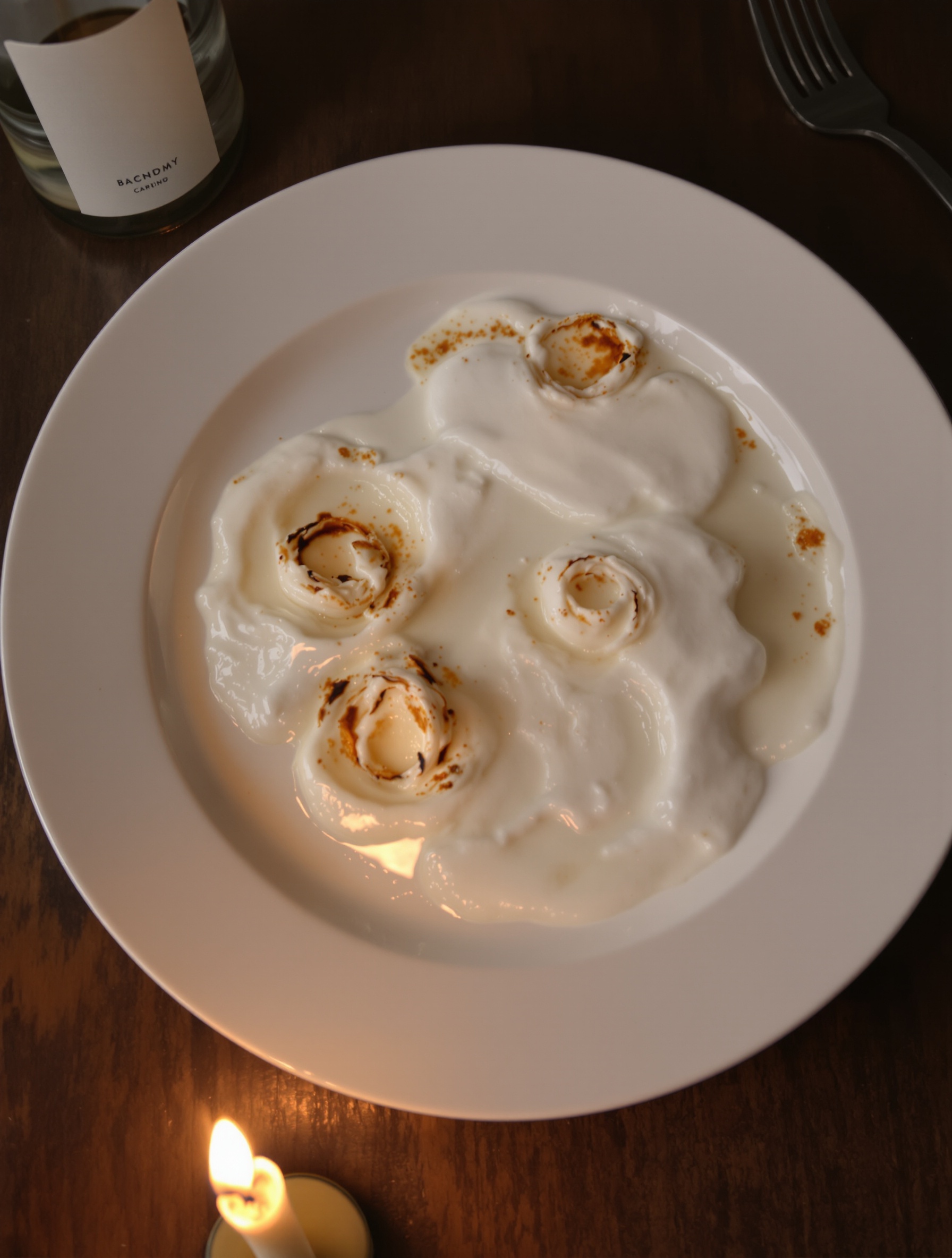 Three spent birthday candles collapsed in hardened wax pools on a white ceramic plate, the last candle flame just out of frame casting a faint warm glow across a dark wooden table.