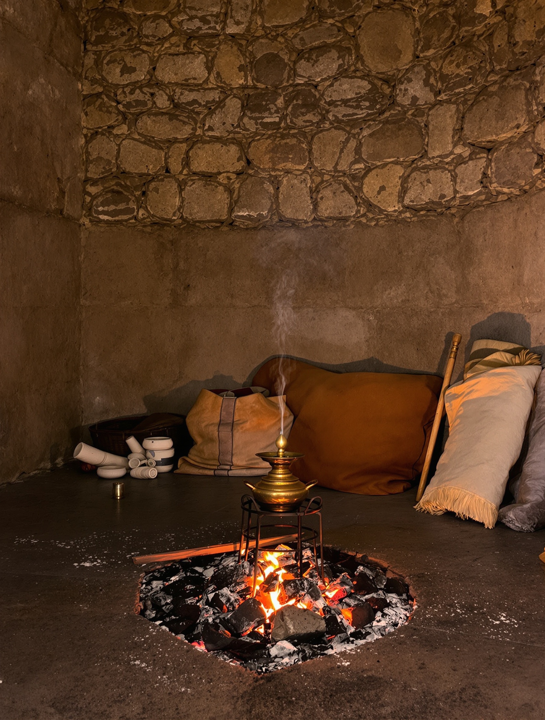 A dim stone interior in Nizwa with a brass qahwa pot smoking over dying embers, evoking the smoldering hospitality of Oman Luxury Caden.