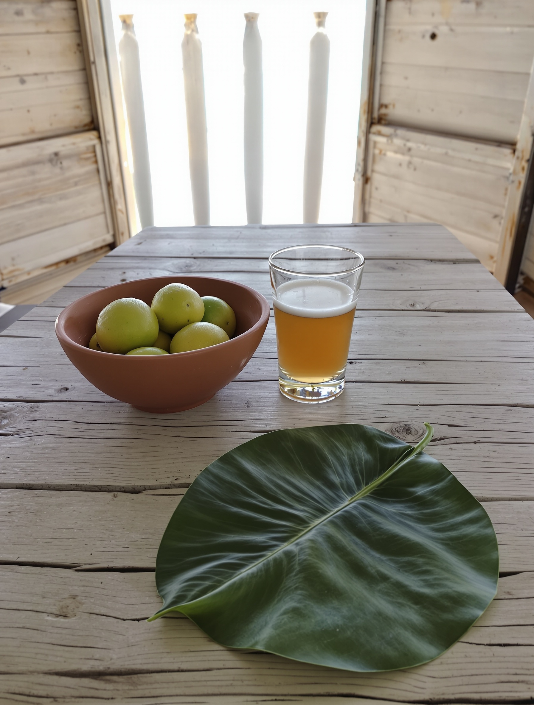 A weathered driftwood veranda table holds fresh limes in a clay bowl, a halved guava, and a pale amber drink, with turquoise ocean softly blurred beyond the railing.