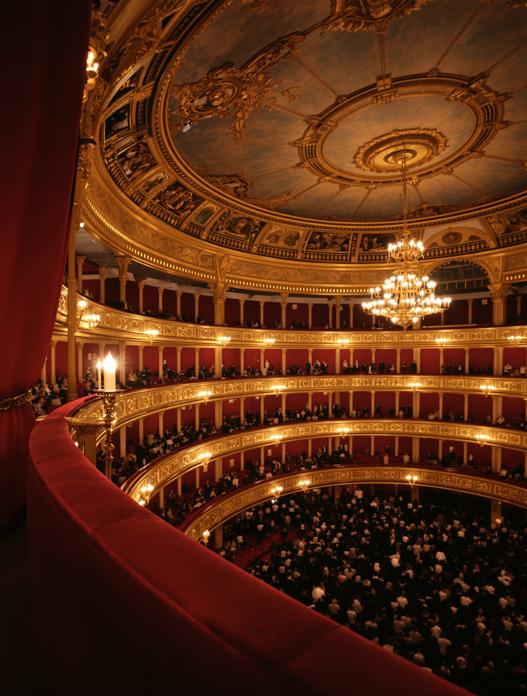 A real editorial photograph inside a gaslit Victorian opera house, shot from a shadowed private box looking across tarnished gilded tiers toward a hazy chandelier, the worn velvet rail in foreground.