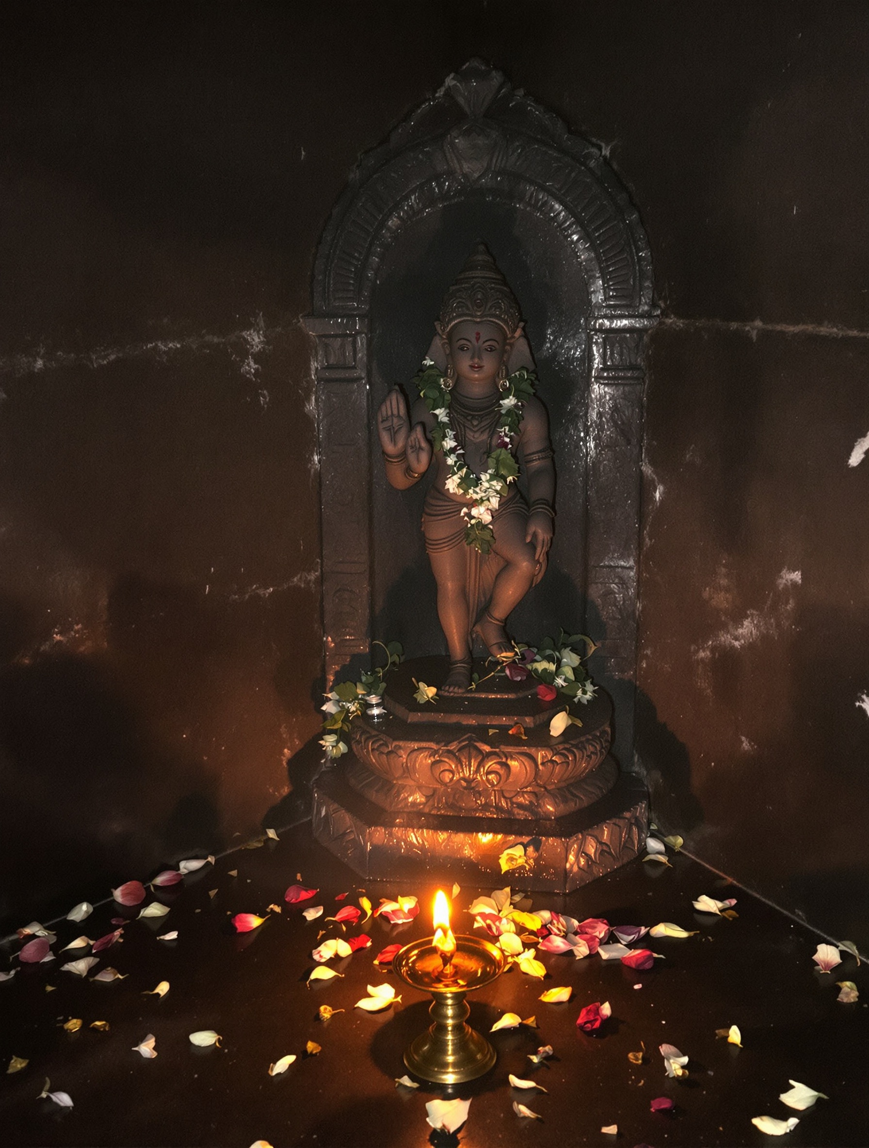 A soot-darkened stone deity idol ringed by wilting jasmine offerings in a dim temple antechamber, evoking the devotional core of Oud Sultani Kannan Koh.