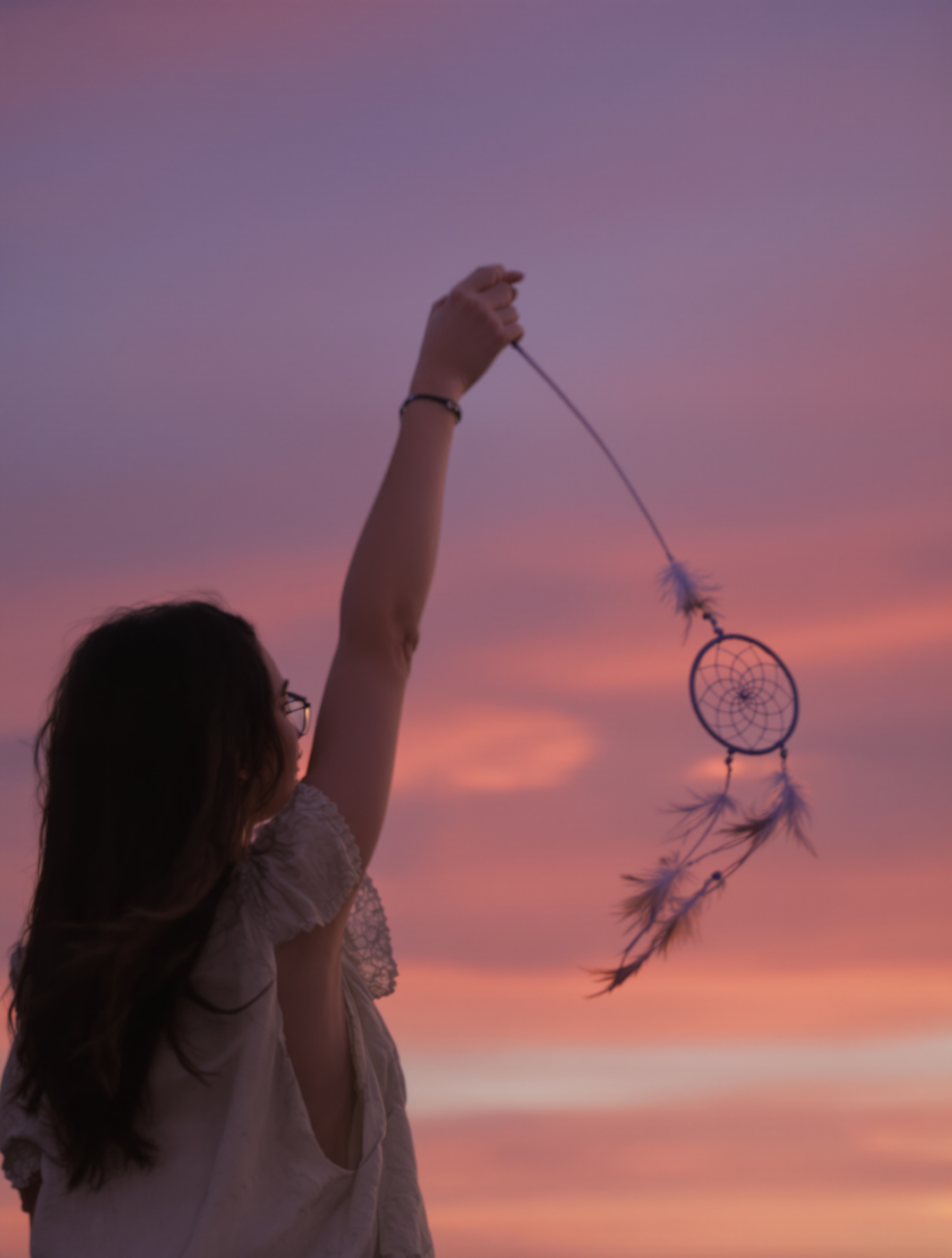 A woman at twilight holds a drifting dreamcatcher at arm's length against a violet-to-amber sky, her back to camera, feathers trailing in the breeze as if moments from release.