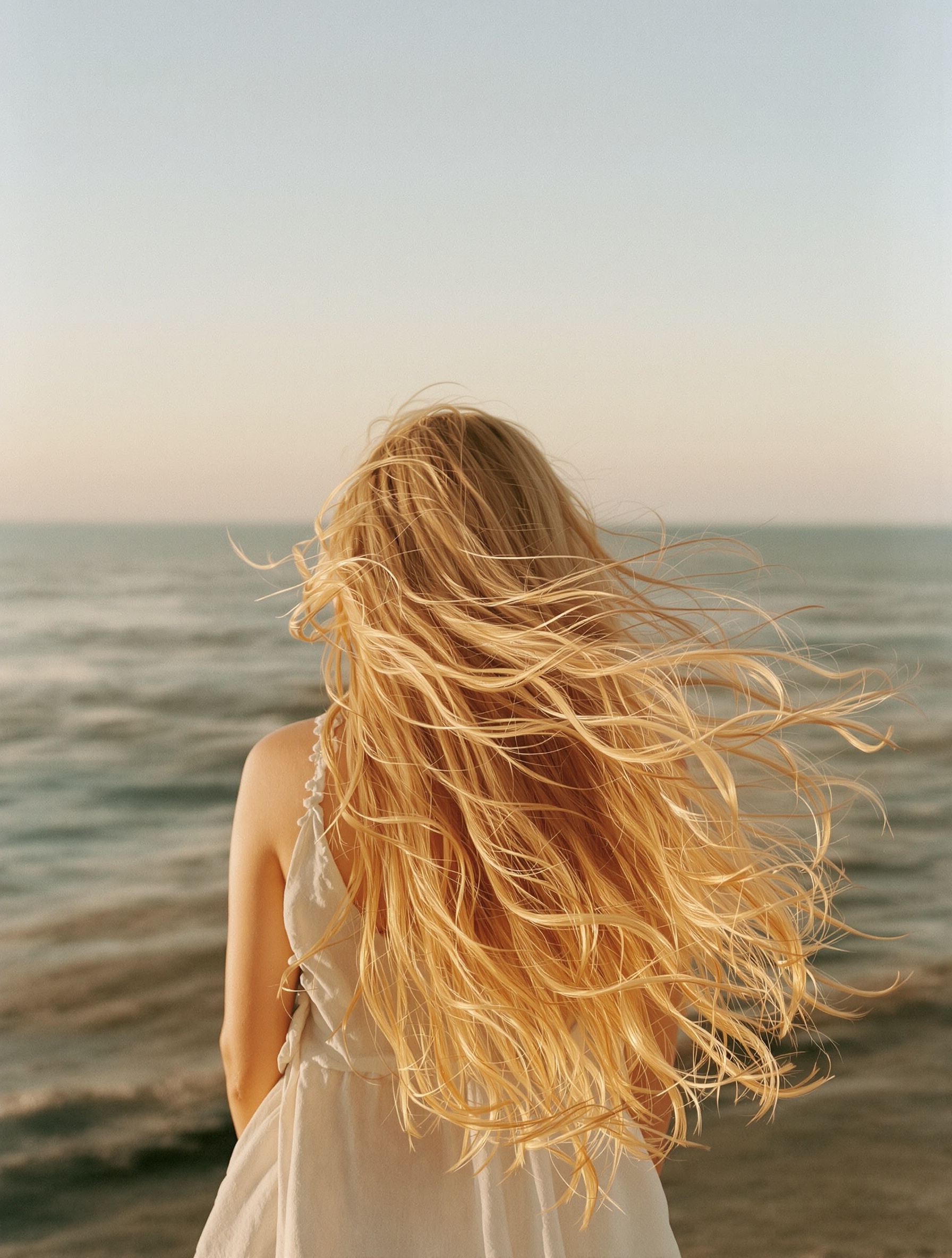 A woman photographed from behind at the shoreline, her long golden hair wind-lifted and glowing against a soft peach and sea-foam dawn sky.