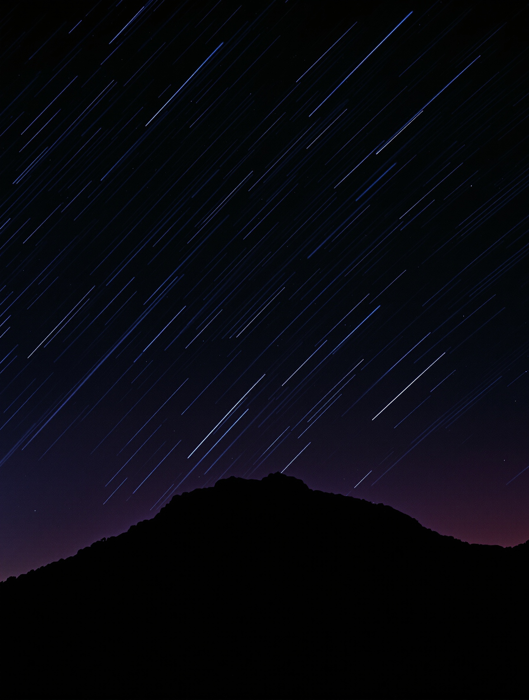 Zdjęcie z długą ekspozycją prawdziwego deszczu meteorów znaczącego srebrzysto-białe smugi na głębokim fioletowoczarnym nocnym niebie ponad ciemną, milczącą sylwetą gór.