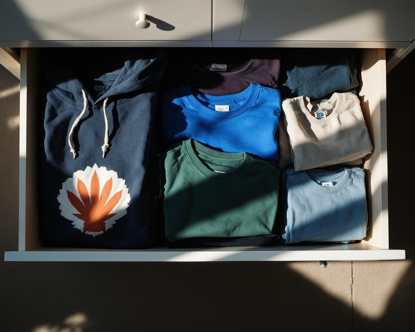 Overhead view of an open wardrobe drawer with neatly folded graphic tees and hoodies, morning light casting soft shadows