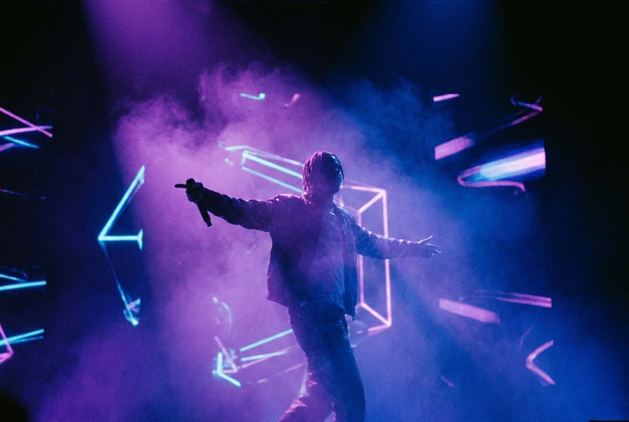 alt text: A colorful music video still showing a singer performing on a neon-lit stage, blending realistic and fantastical AI-generated visual elements like floating geometric shapes and surreal light trails