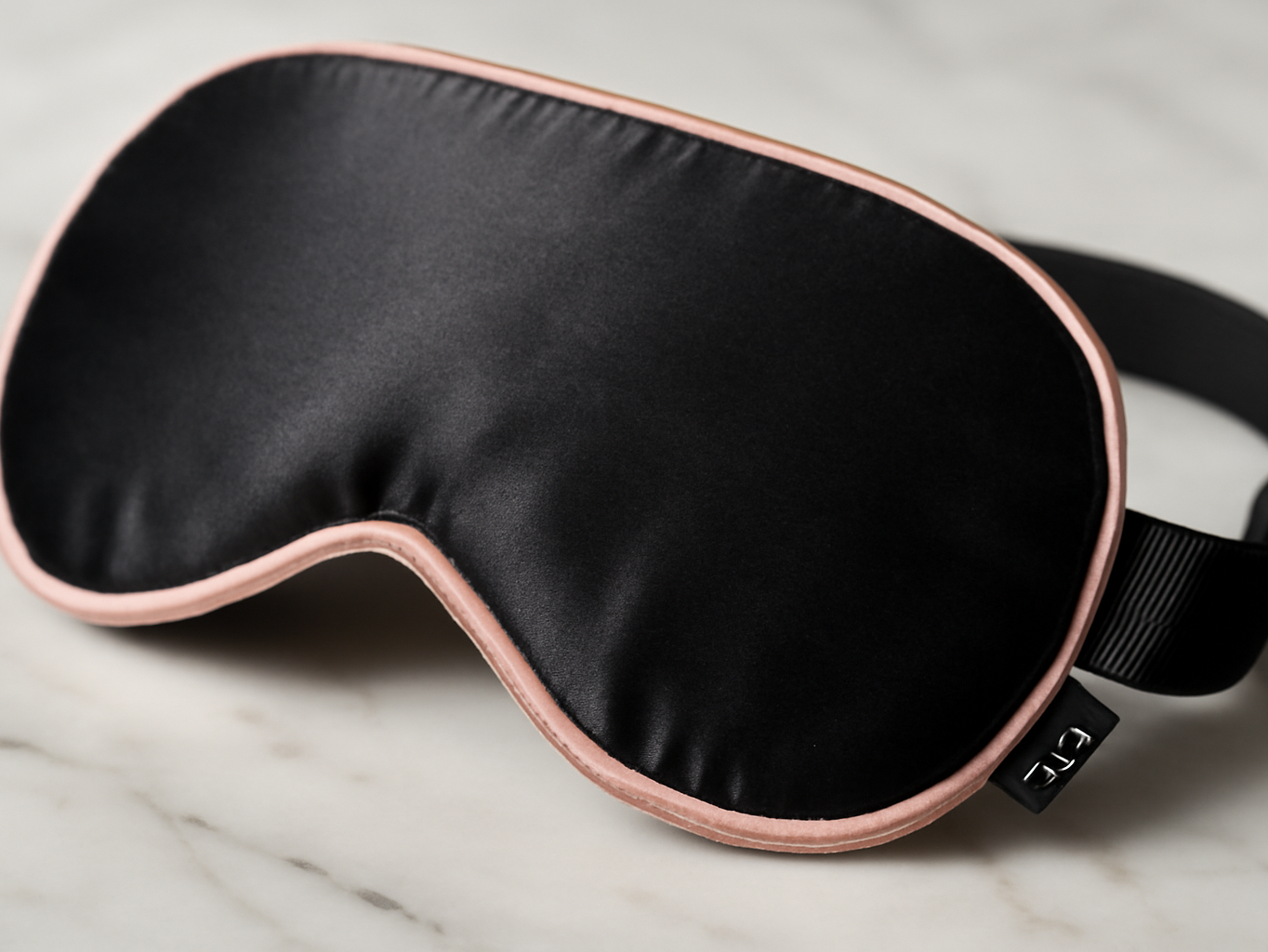 Silk flat sleep eye mask in black with pink trim, macro close-up showing smooth fabric texture on marble